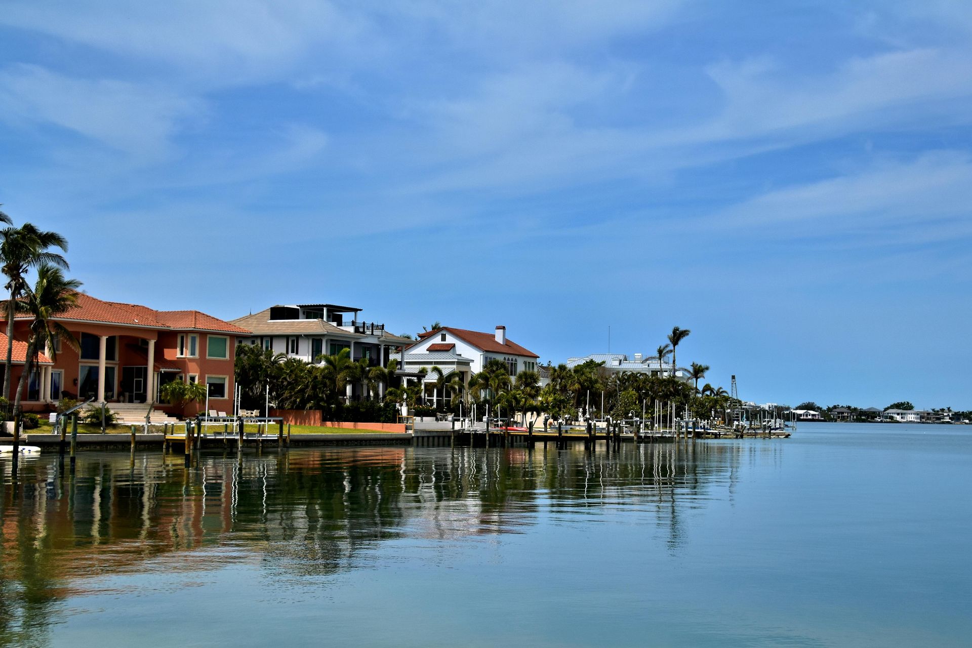 Luxurious waterfront houses sit along a calm canal under a bright blue sky, with palm trees and reflections on the water.