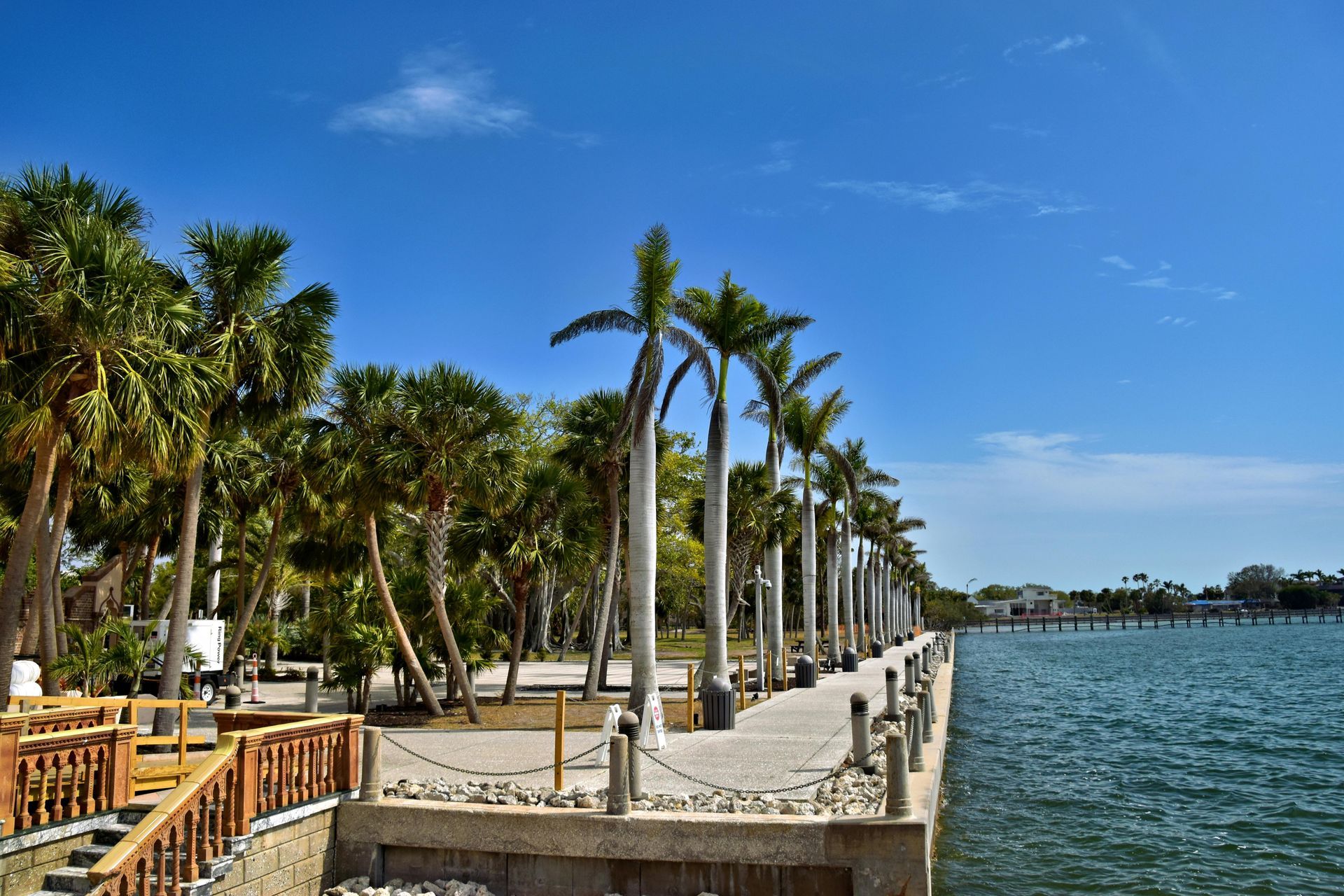 A paved walkway lined with tall palm trees runs along a calm, blue waterfront under a bright, sunny sky.