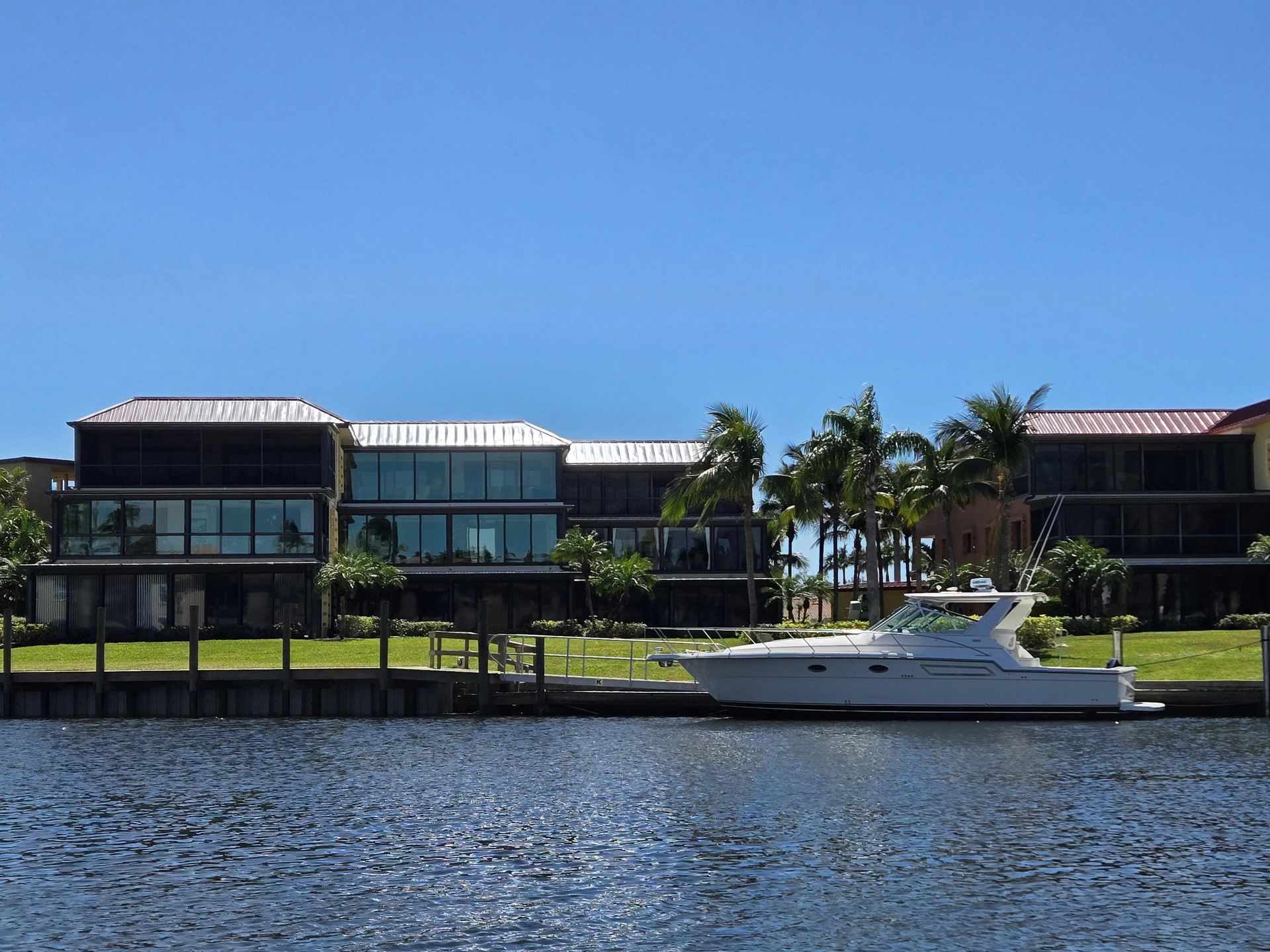 A white boat docked in a canal in front of modern, multi-story waterfront houses under a clear blue sky.