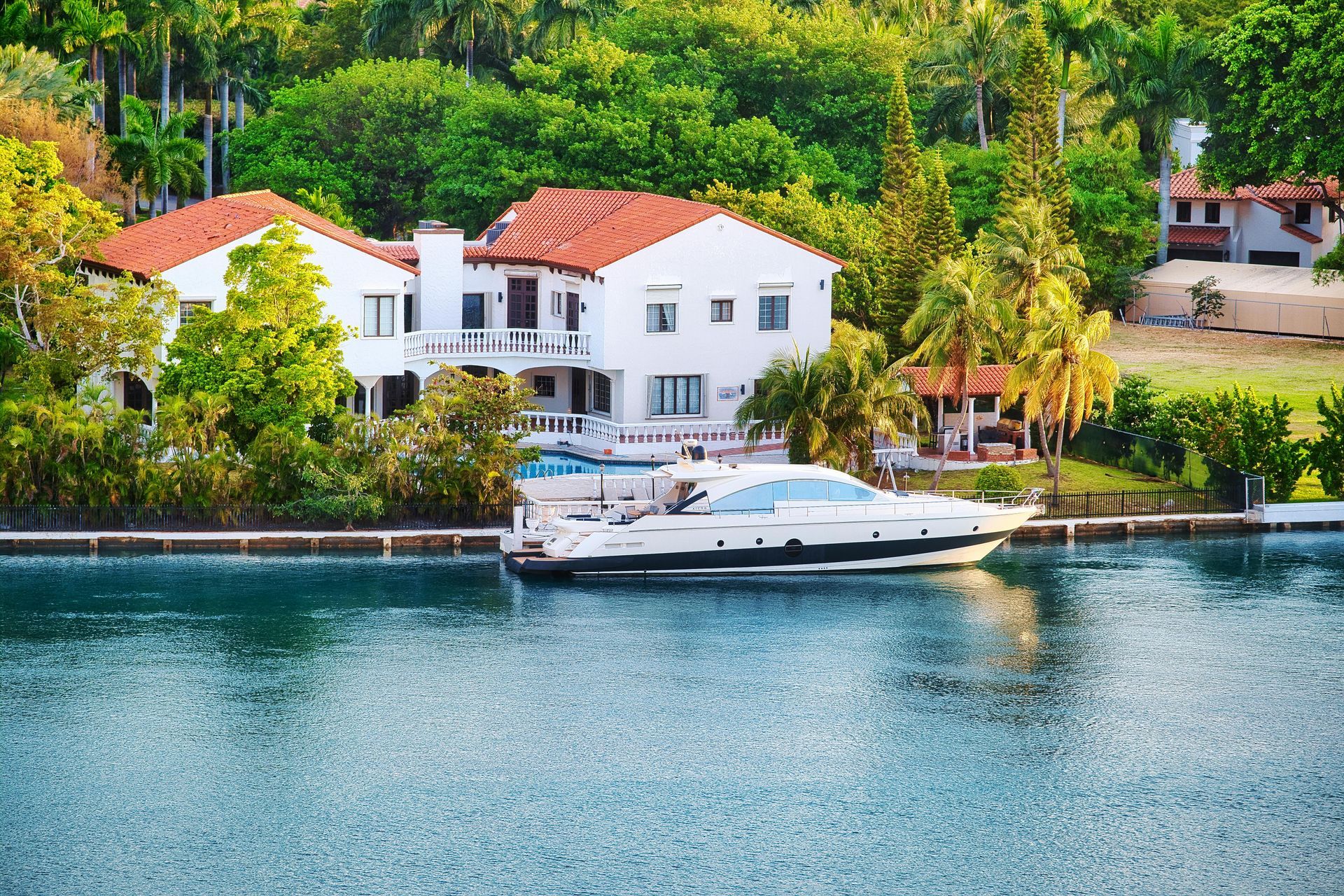 A white yacht is moored in a tranquil canal beside a multi-story white house with a red tiled roof, surrounded by trees.