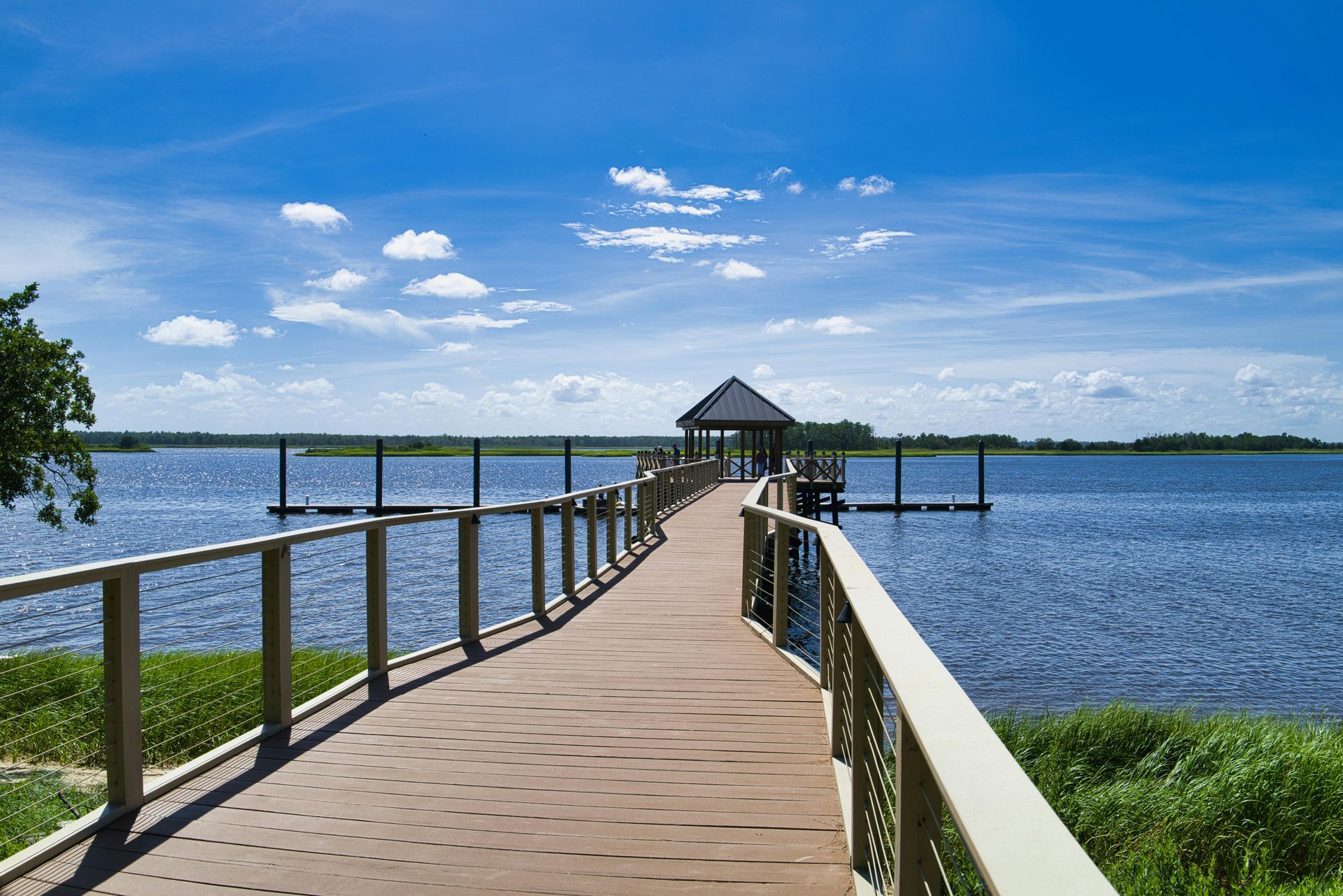 A wooden pier extends over a calm blue lake toward a small covered gazebo under a bright, sunny sky.