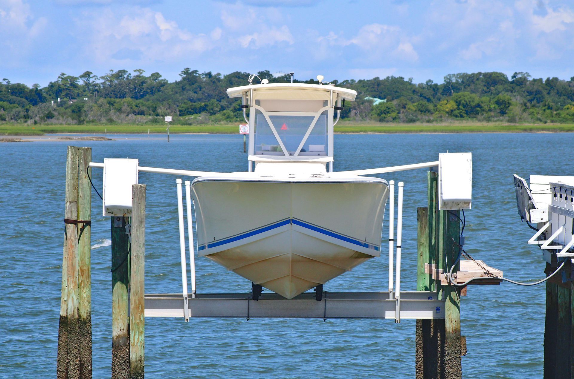 A white boat suspended on a mechanical lift above calm blue water with a marshy shoreline in the background.