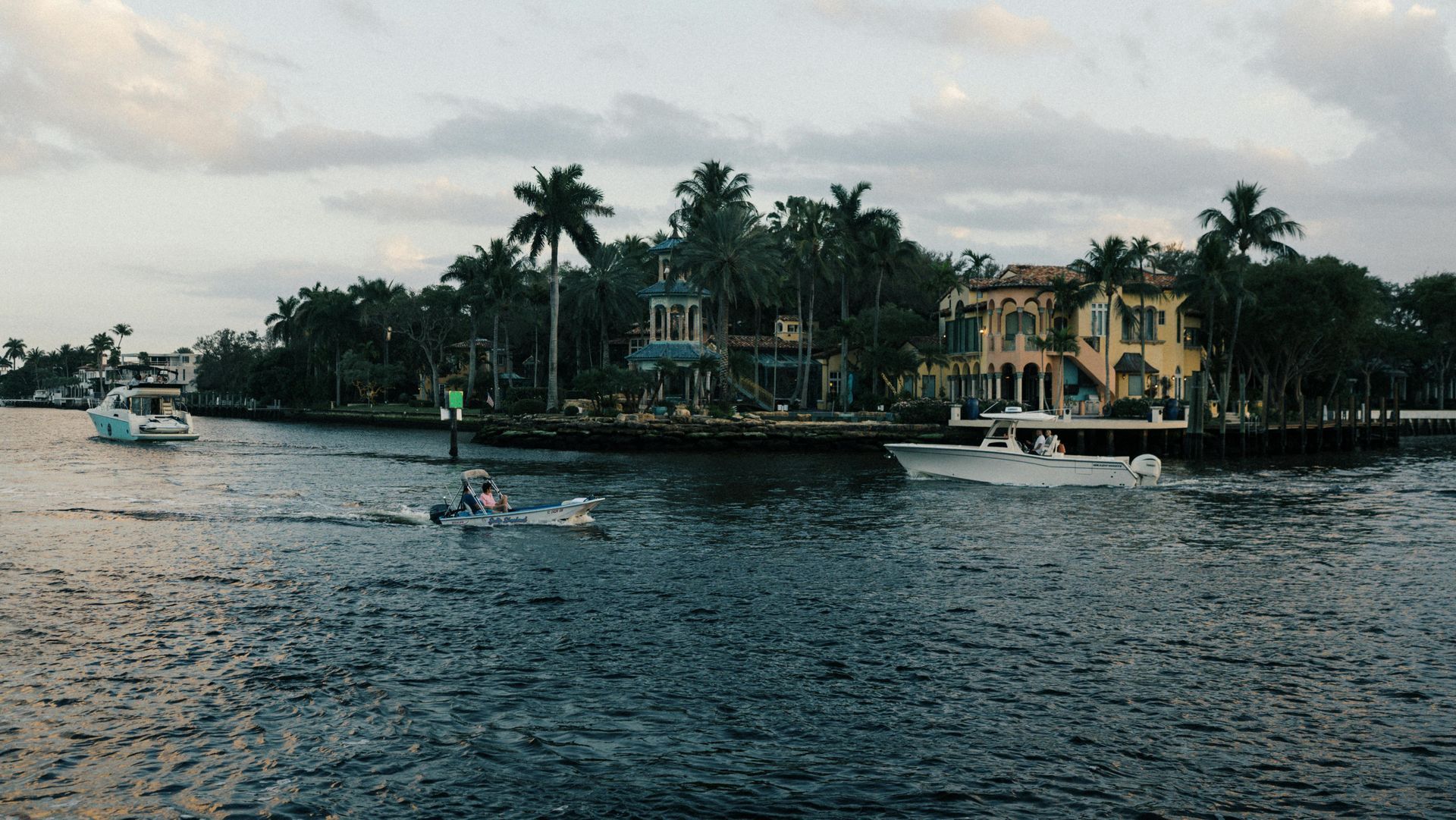 Boats move along a calm canal past a large Mediterranean-style waterfront house surrounded by palm trees at dusk.