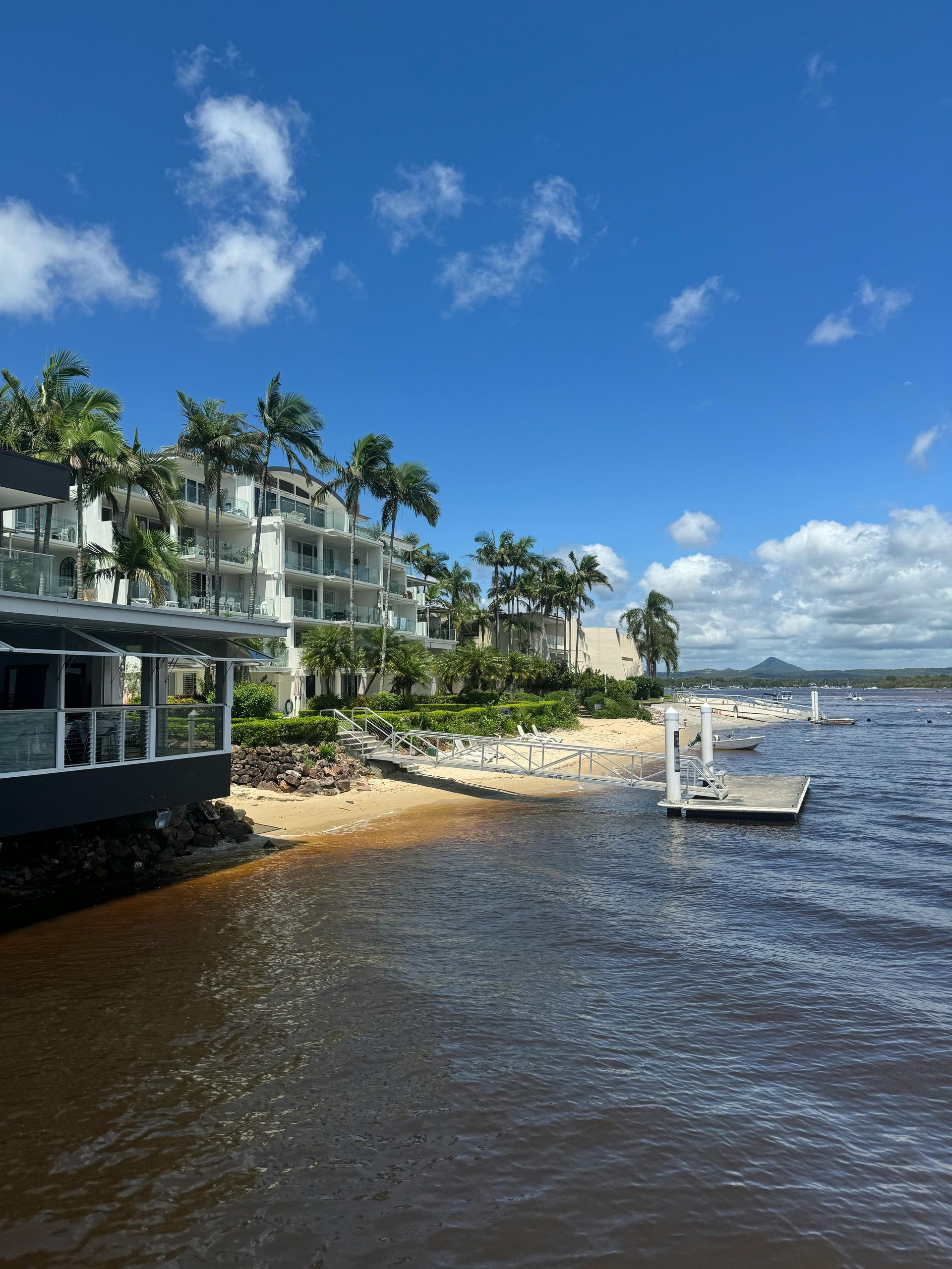 A sunny waterfront view of a multi-story hotel resort with palm trees, a sandy beach, and a small wooden dock.