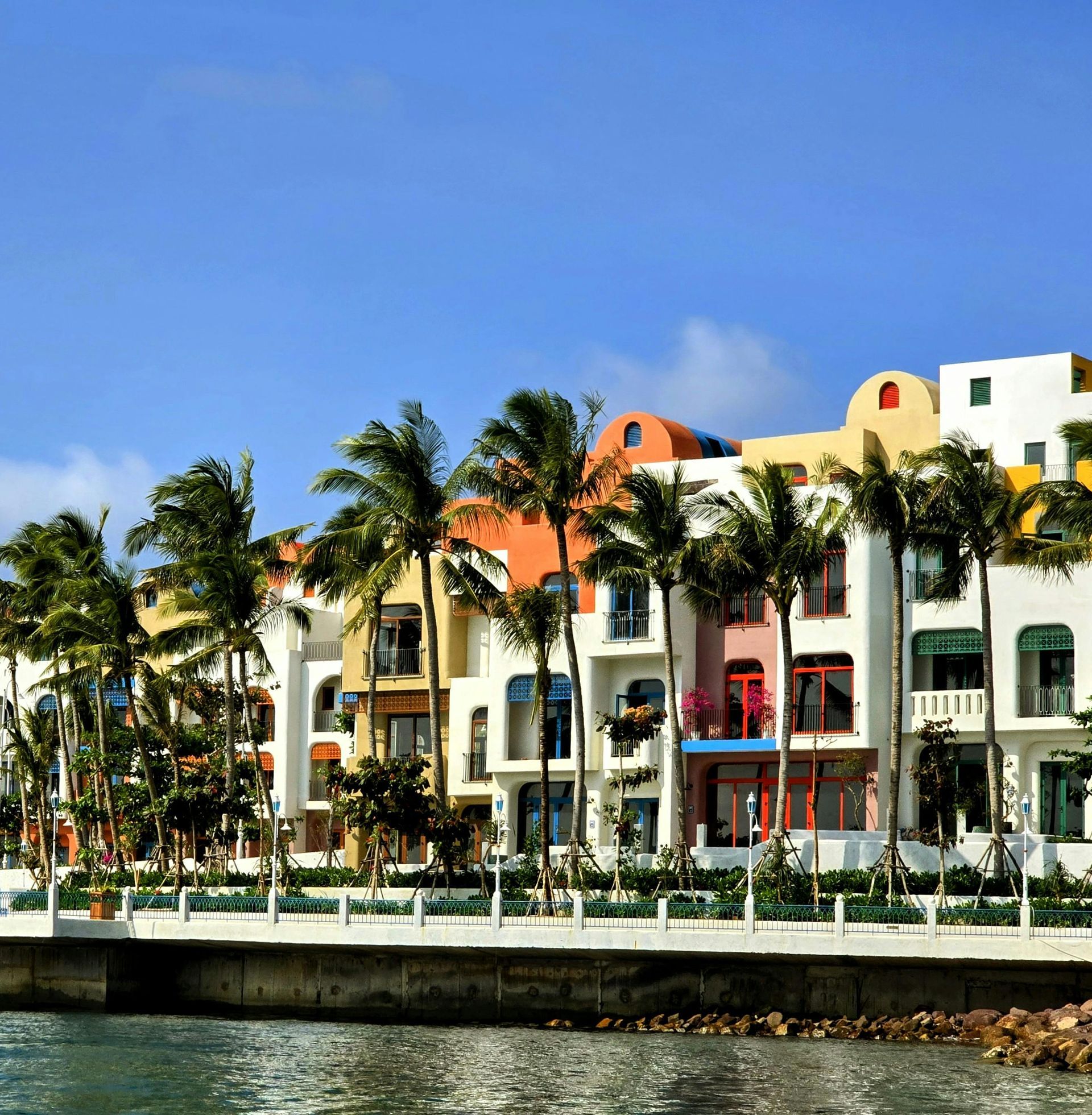A row of colorful, Mediterranean-style buildings lined with palm trees stands along a stone waterfront under a blue sky.