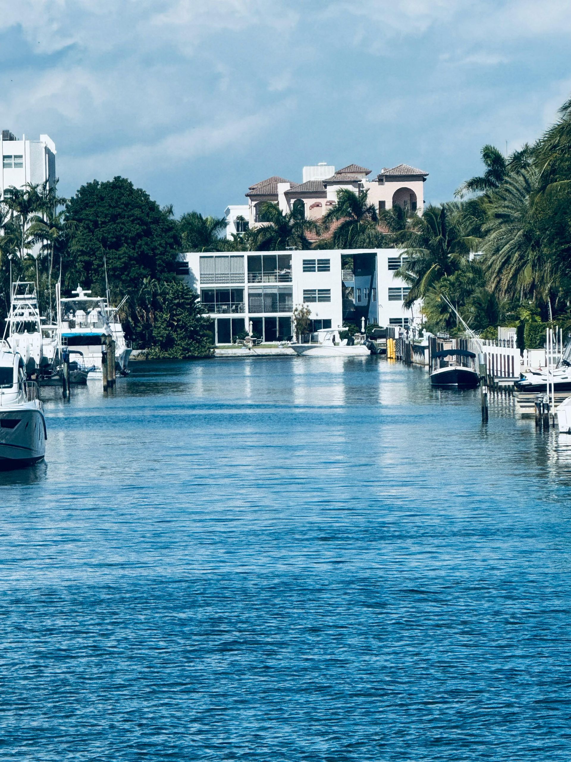 A narrow canal with yachts moored along the sides, leading to a modern white residential building under a blue sky.