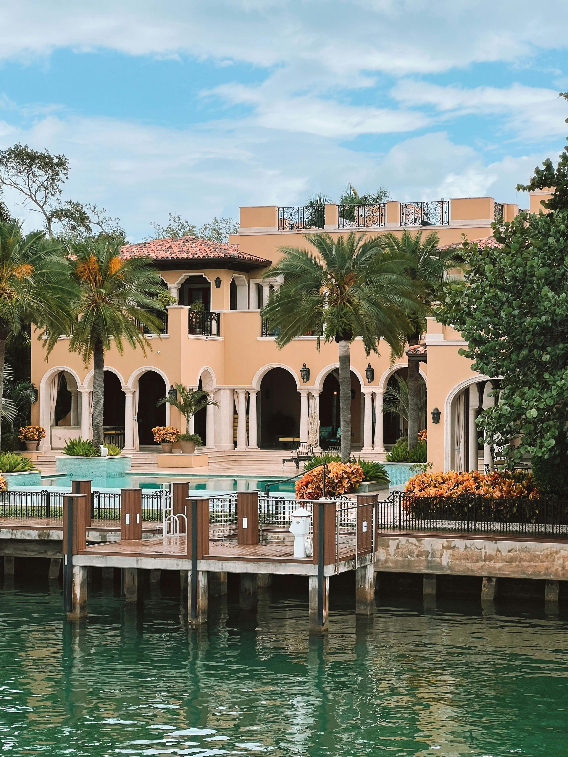 A beige, Mediterranean-style villa with arched doorways overlooks a calm waterway from a wooden dock.