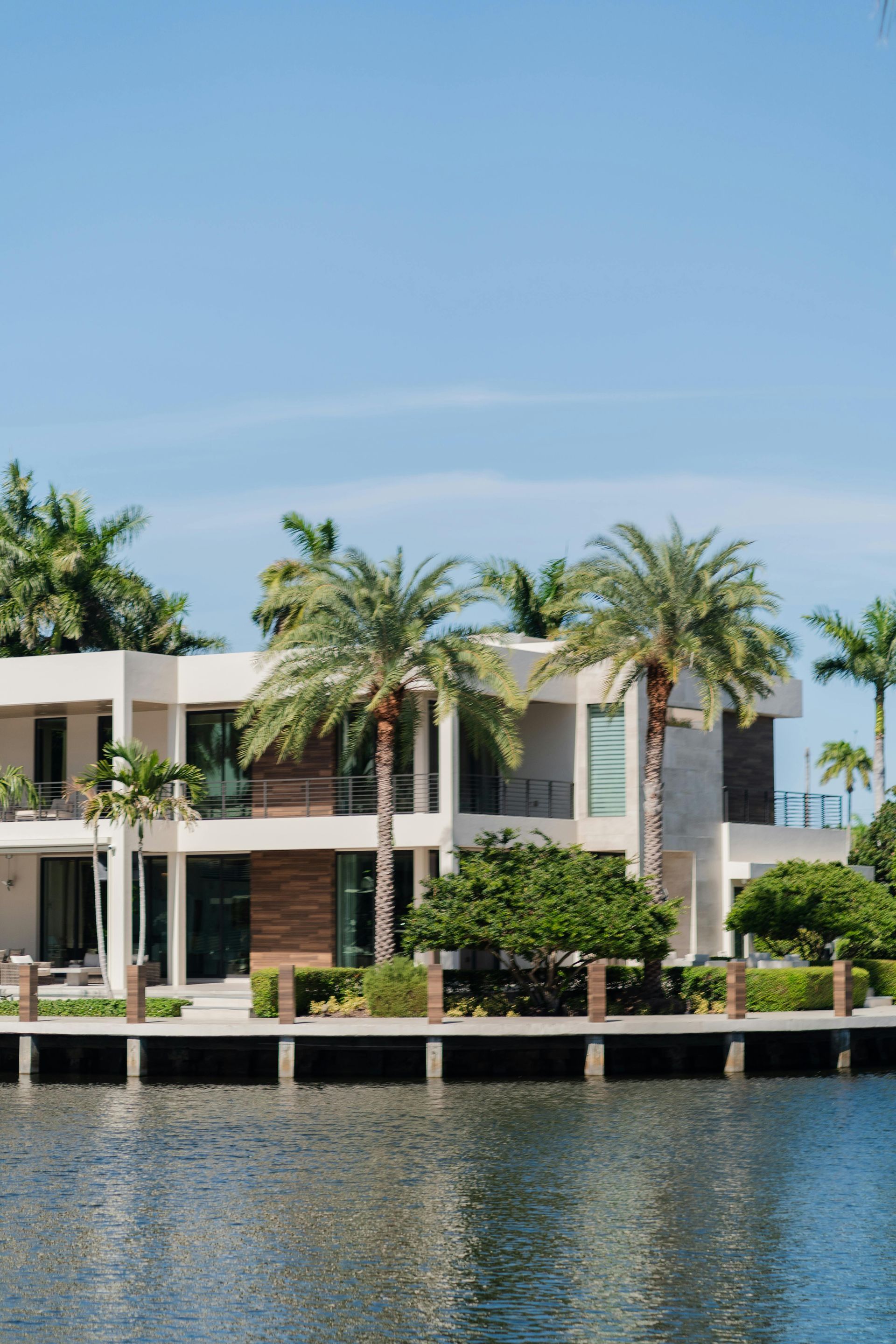 A modern, two-story waterfront house with beige walls and wood accents, surrounded by palm trees, beside a calm canal.