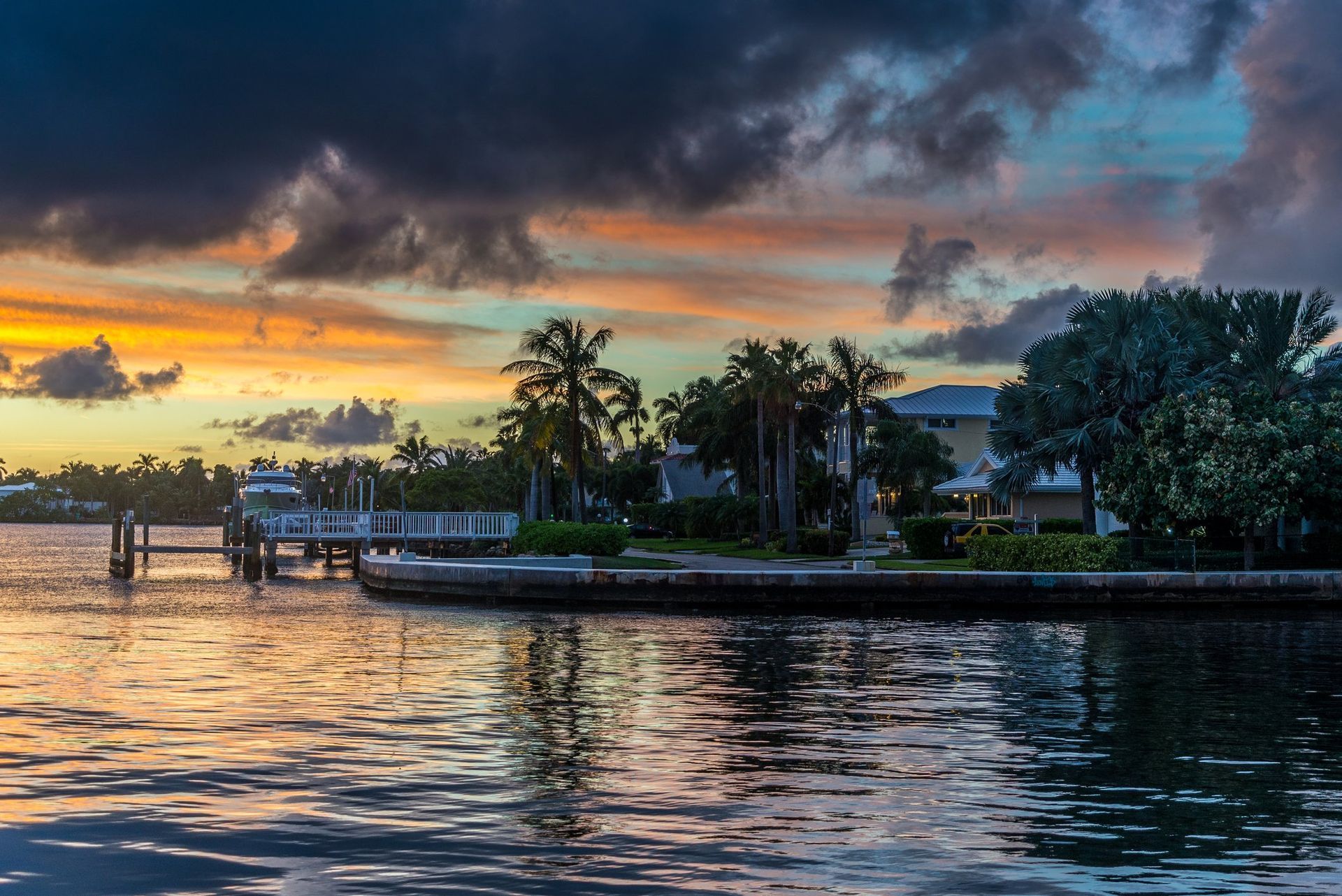 A scenic sunset with orange and purple clouds over a waterfront property, palm trees, and a wooden pier.