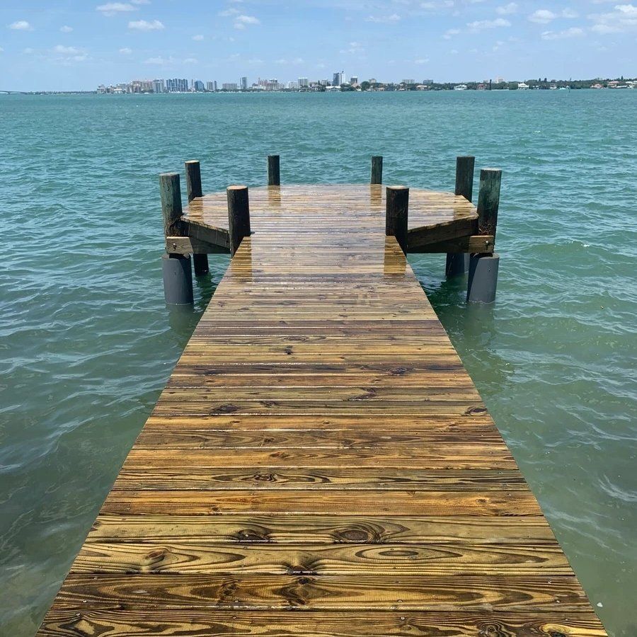 A wooden dock extends into calm, turquoise water toward a distant city skyline under a sunny, partly cloudy sky.