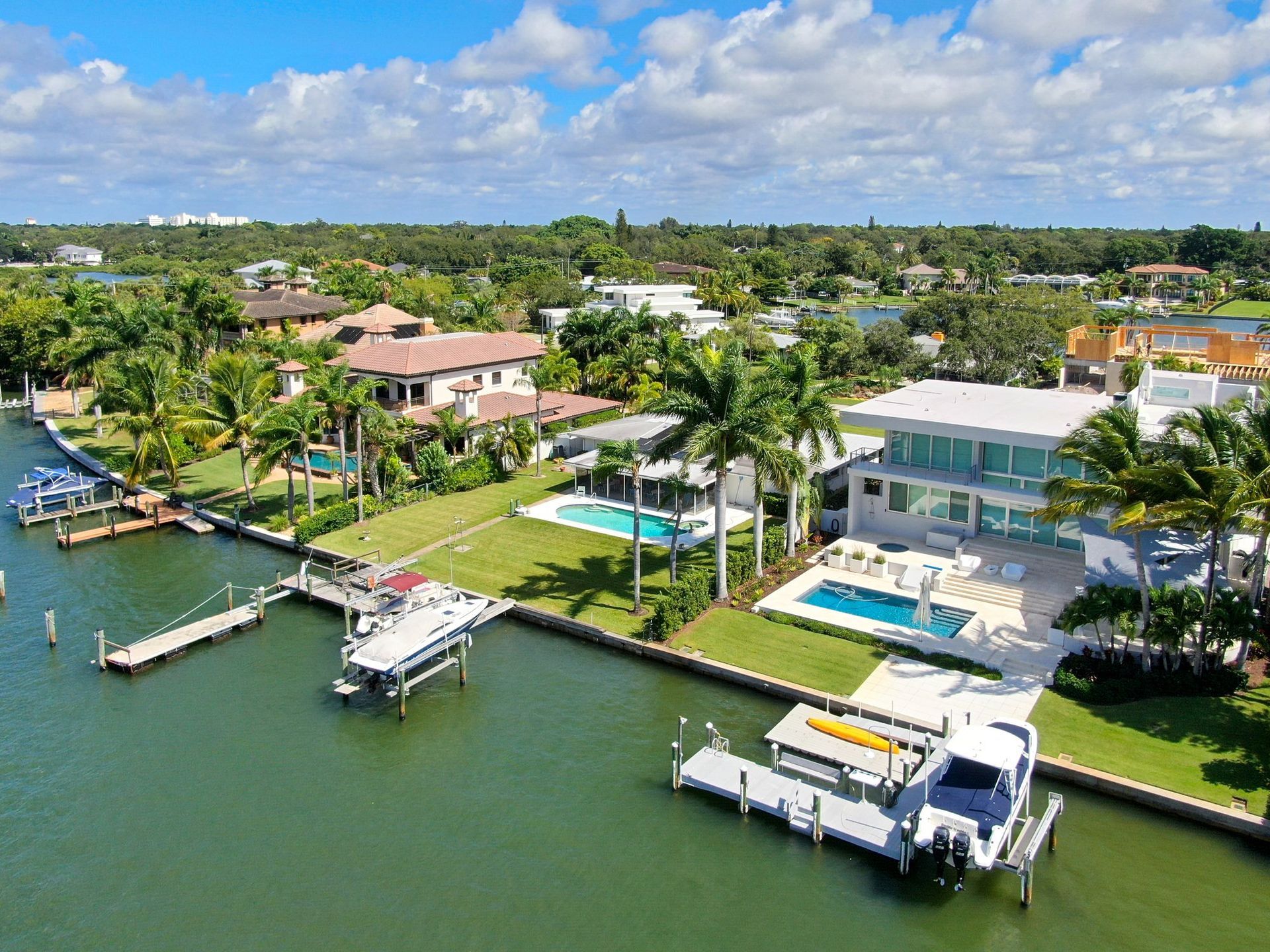 Aerial view of two modern waterfront houses with swimming pools and private docks on a canal, surrounded by palm trees.