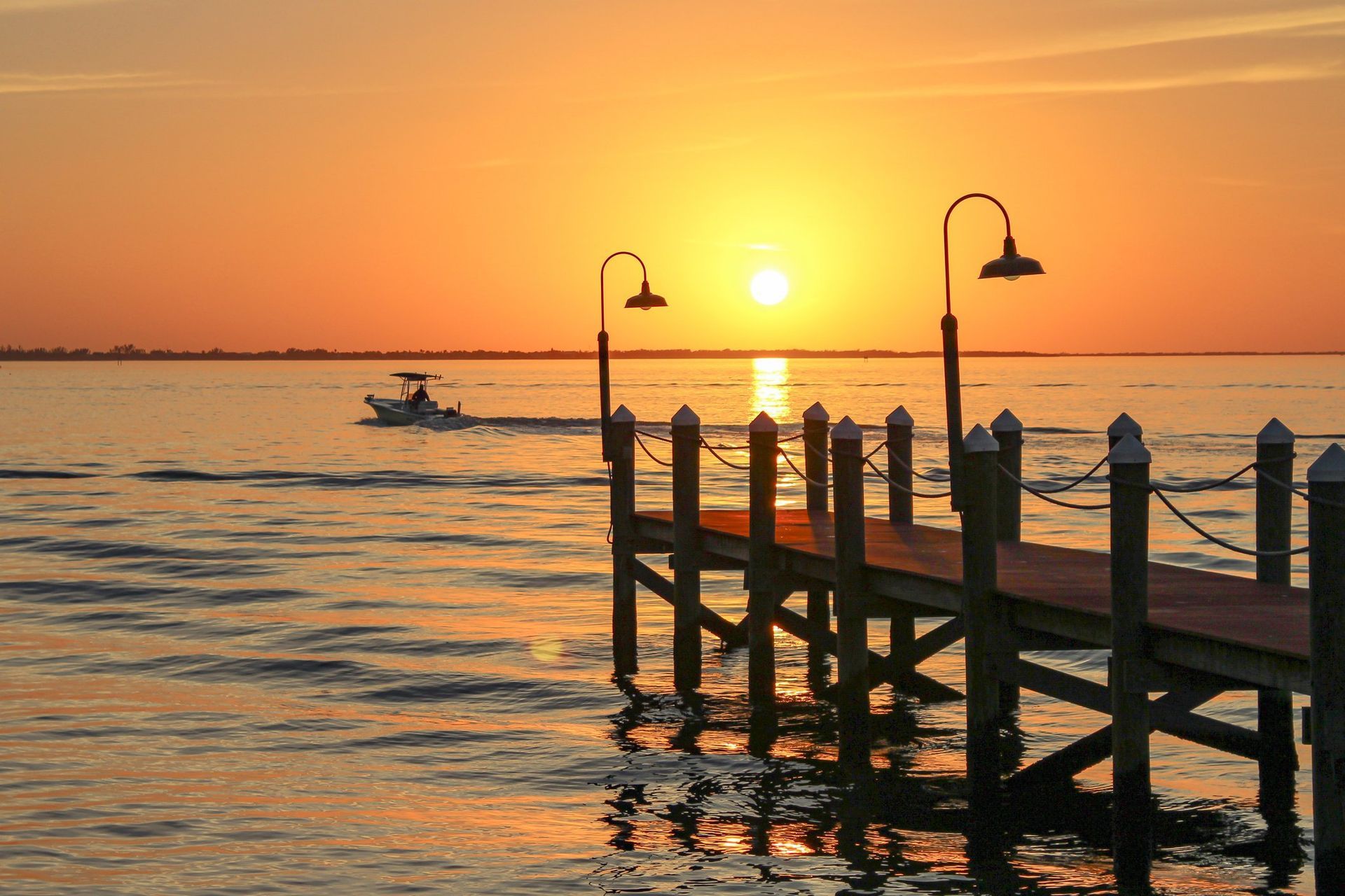 A wooden pier extends into calm water under a golden sunset, with a small boat traveling in the distance.