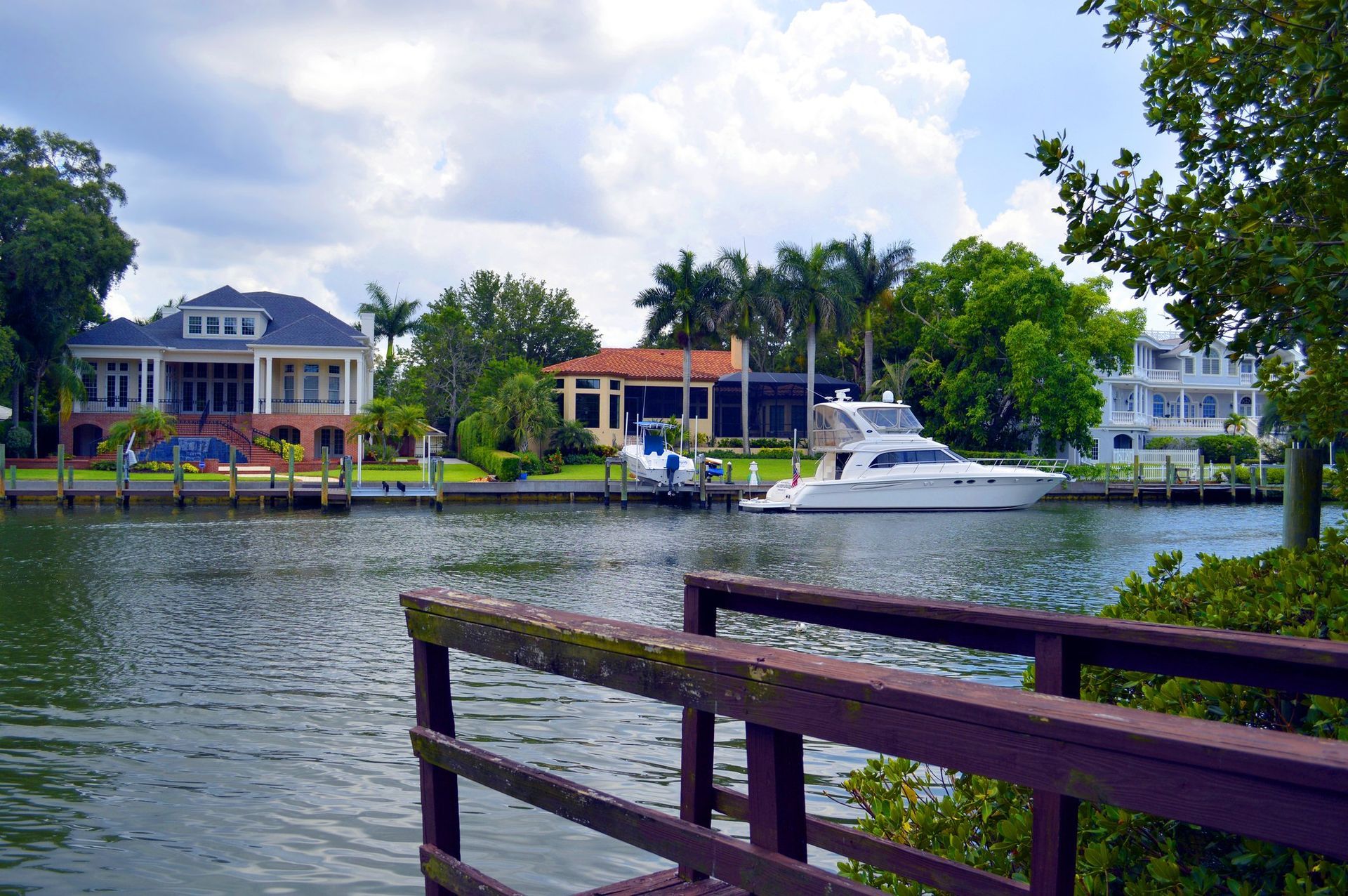 A canal view featuring waterfront homes, a docked motor yacht, and a wooden bridge in the foreground under a cloudy sky.