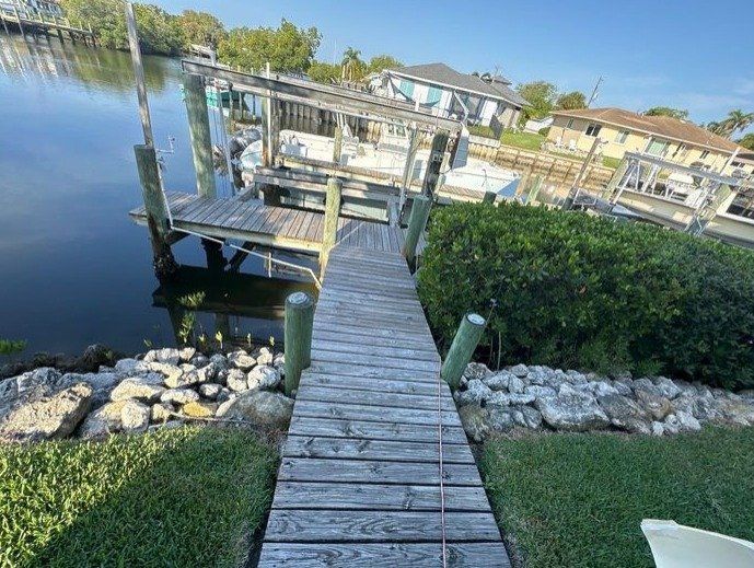 A weathered wooden dock extends from a grassy lawn into a calm canal, leading to a covered boat slip under a clear sky.