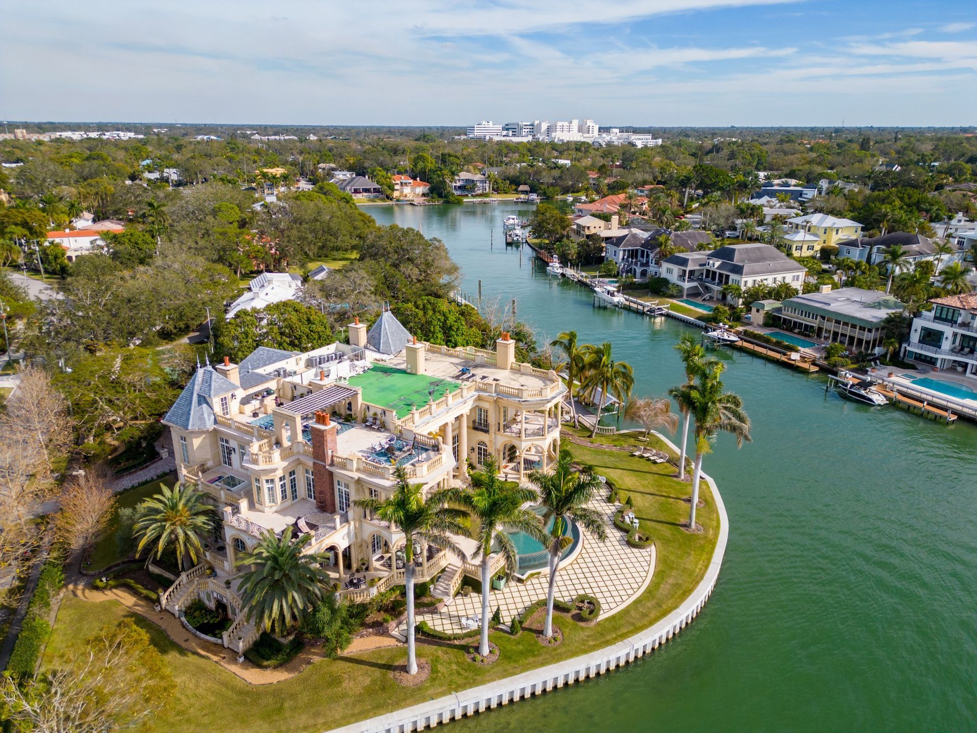 Aerial view of a sprawling, light-colored mansion on a point of land next to a canal with palm trees and a swimming pool.