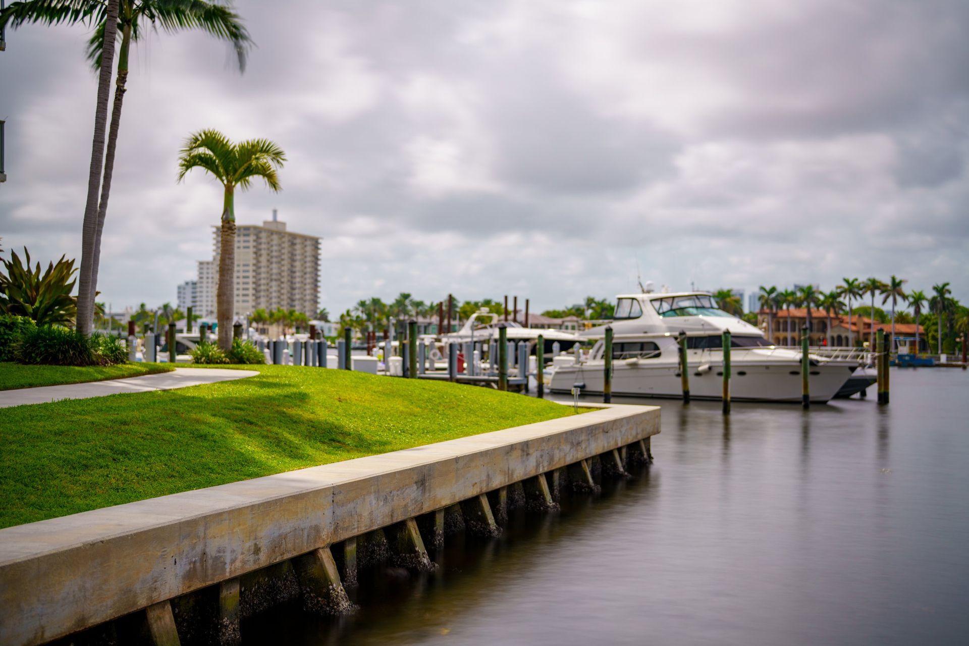 A white yacht docked at a concrete pier next to a grassy lawn with palm trees, under a cloudy sky.