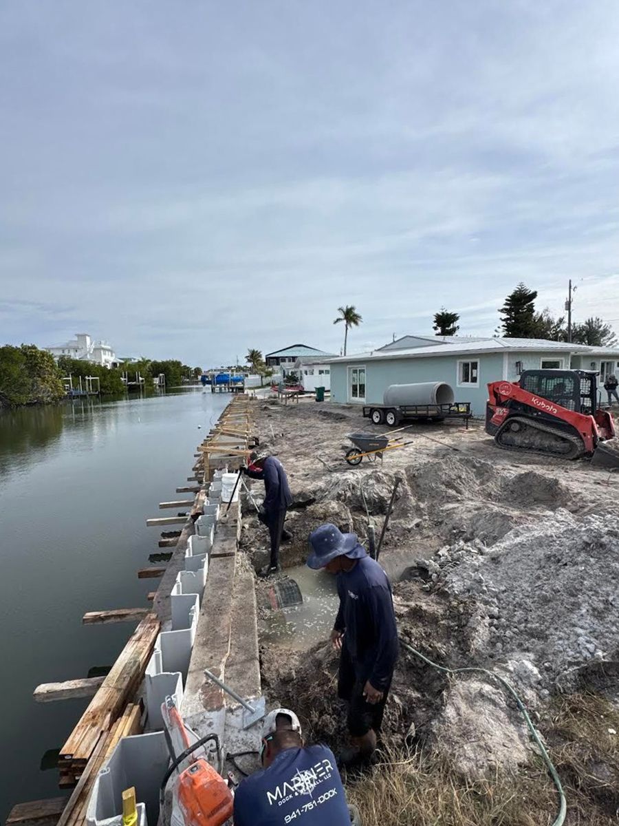 Three workers install a concrete seawall along a canal next to a light-colored house and construction equipment.
