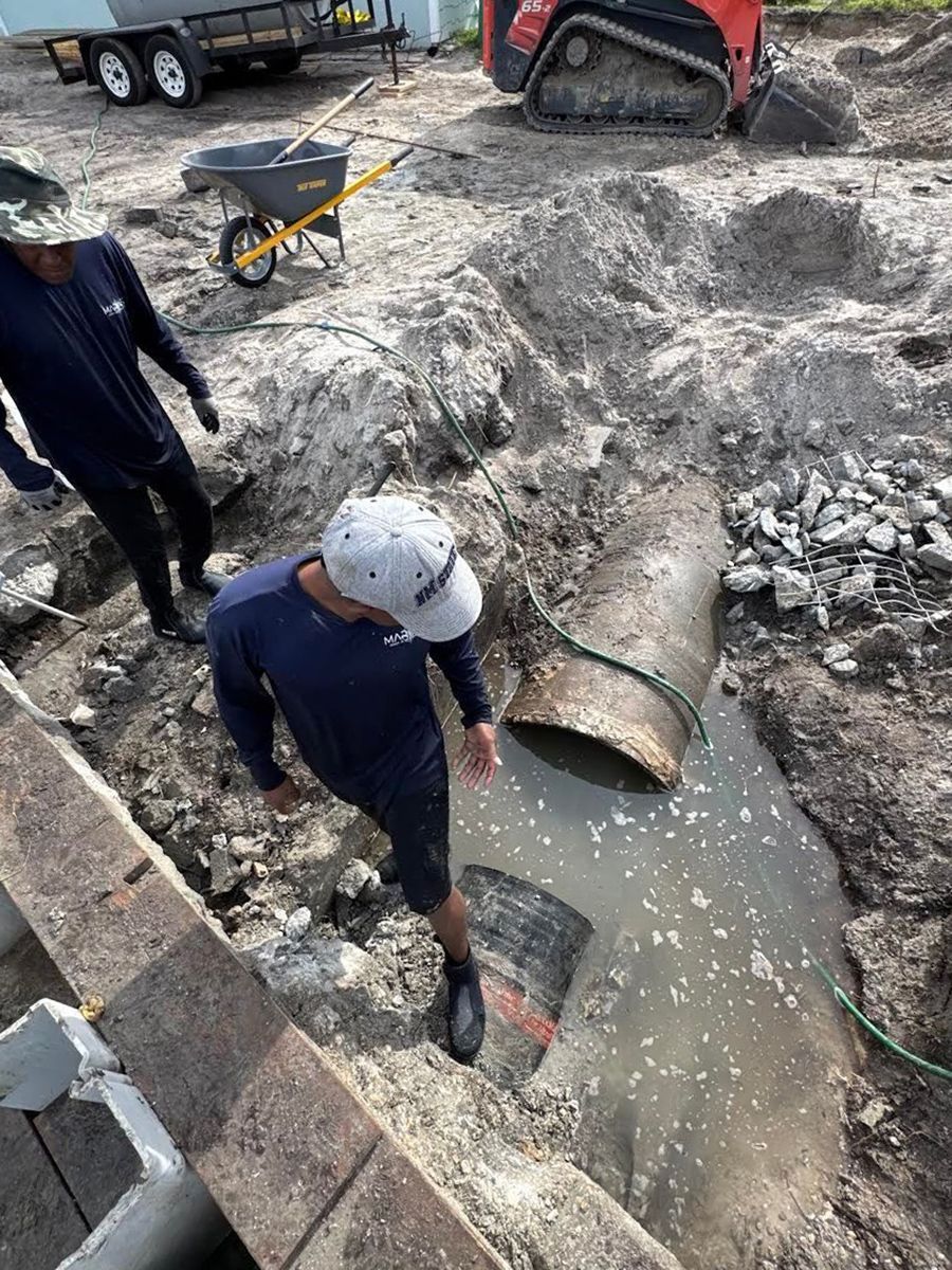 Two workers stand near an excavated trench containing two separated pipe ends in a construction site.