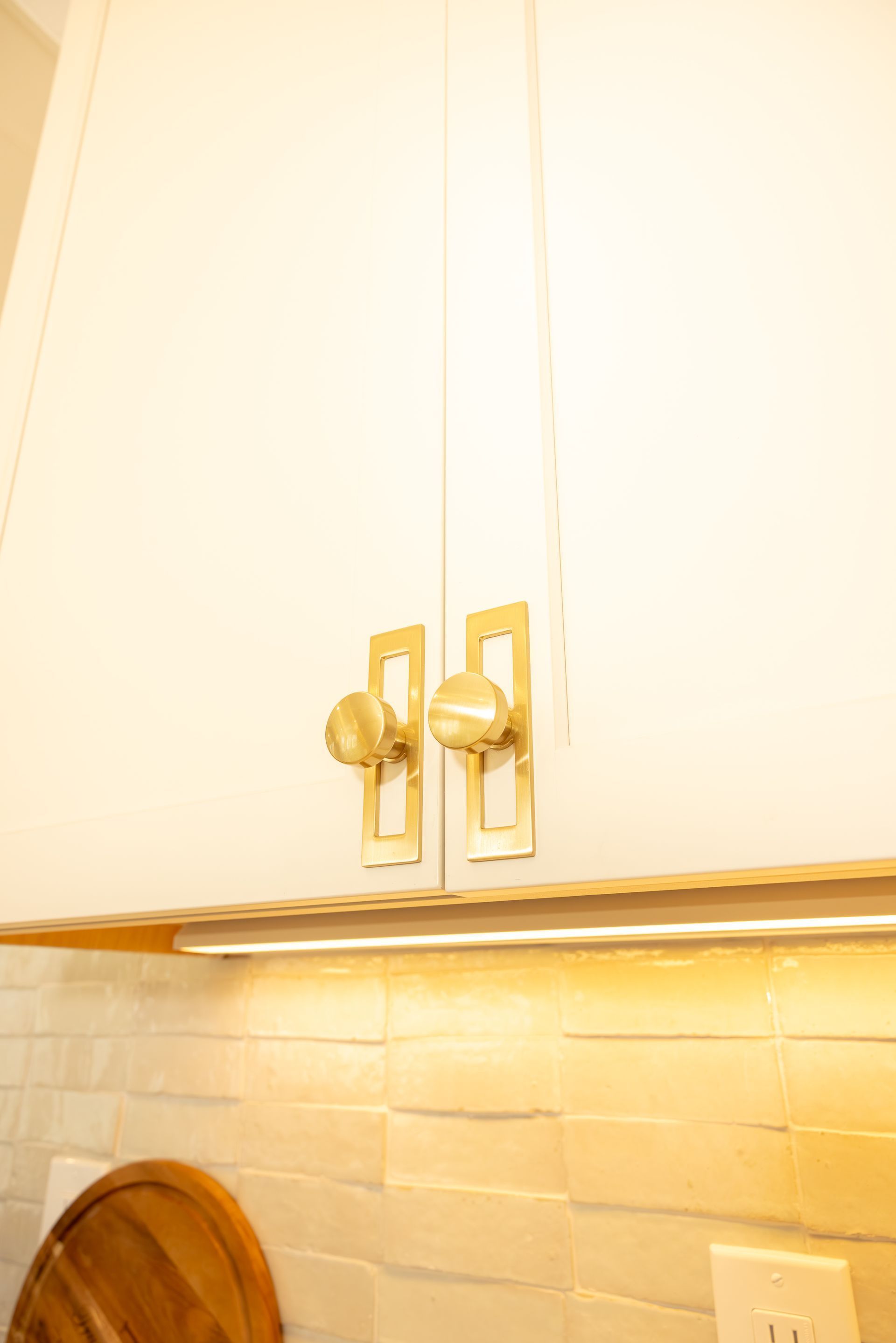 White kitchen cabinets with brass handles above a lit tiled backsplash and wooden countertop edge