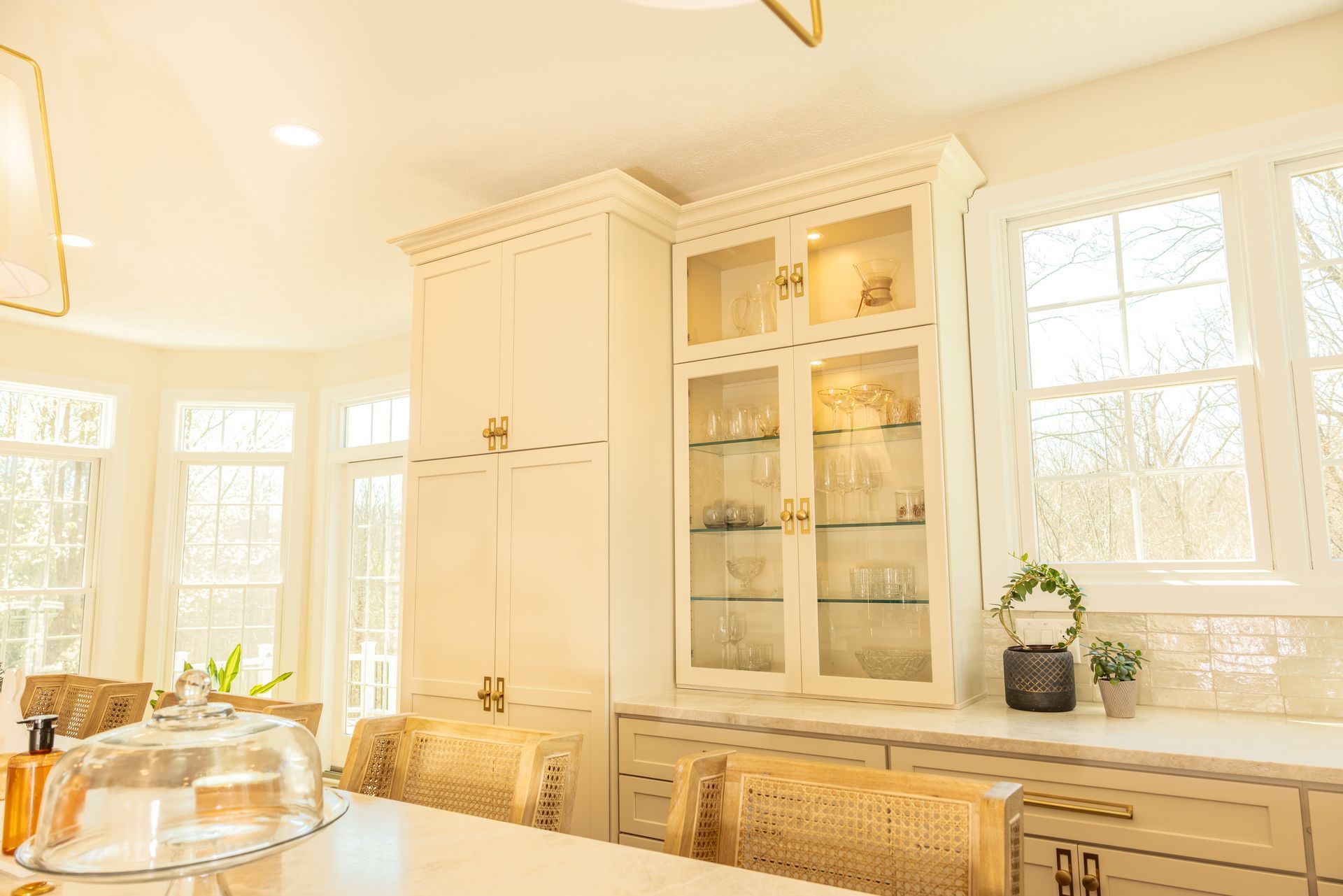 Bright white kitchen with glass cabinets, large windows, and wicker chairs by a dining table