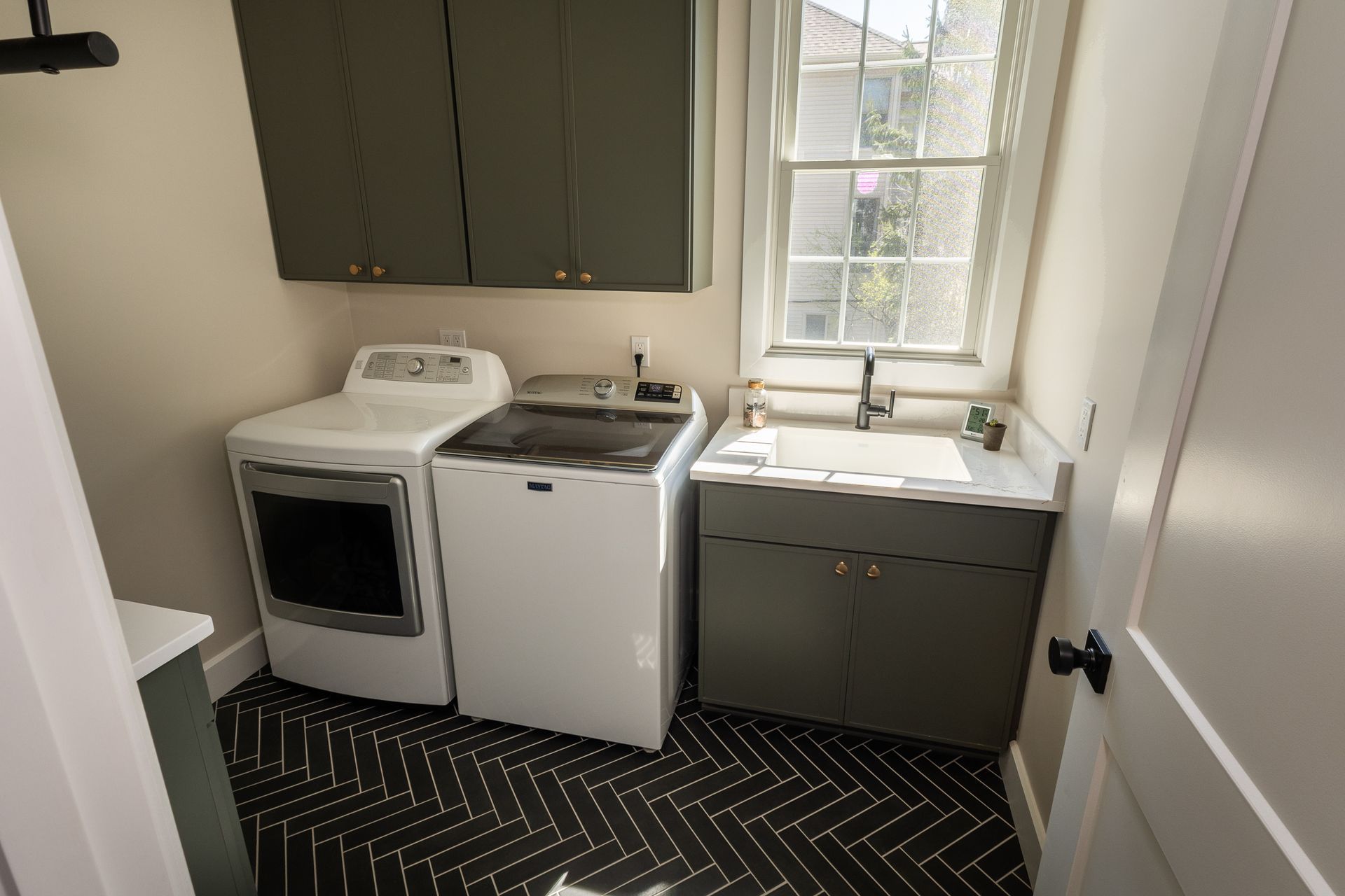 Small kitchen with white appliances, gray cabinets, and a window above the sink on a black patterned floor.