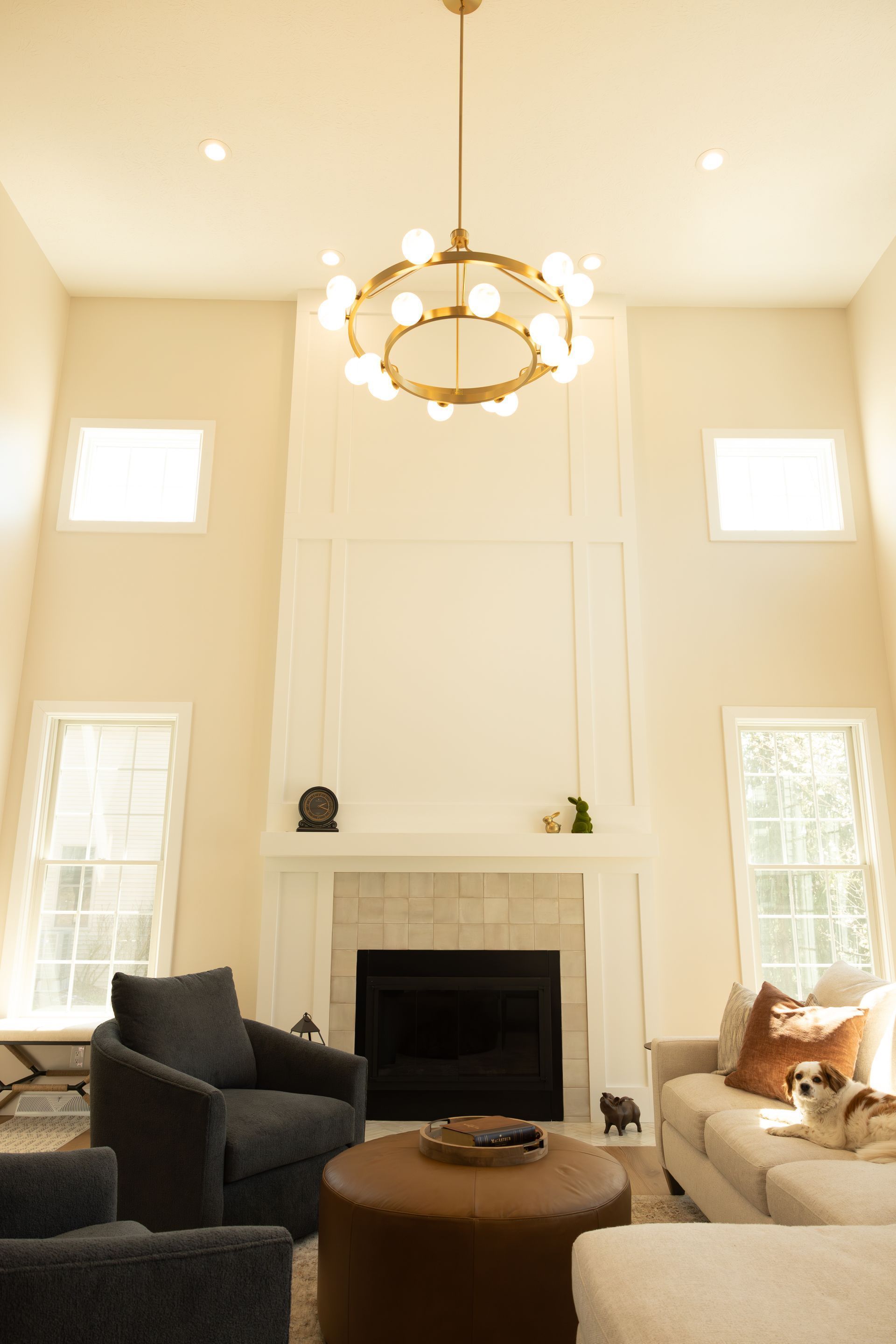 Bright living room with tall windows, white fireplace, chandelier, and dark chairs around a round coffee table