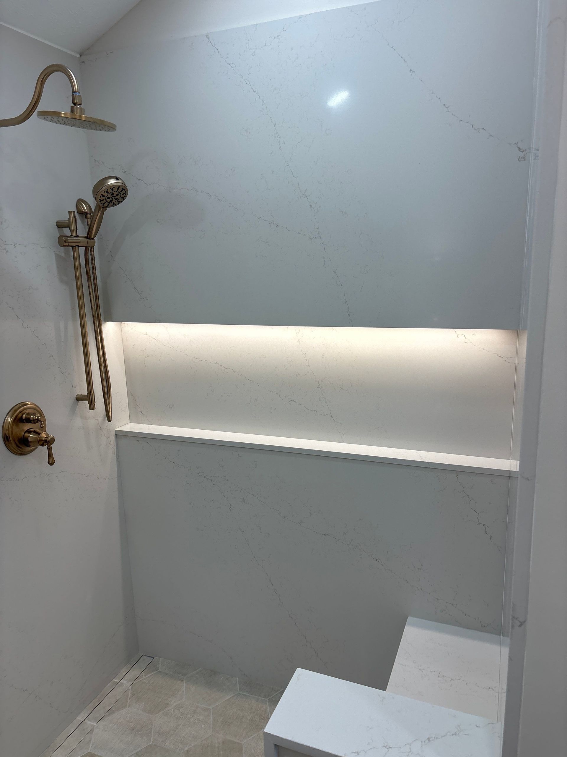 Empty shower stall with white walls, chrome fixtures, built-in shelf, and a small bench or step.