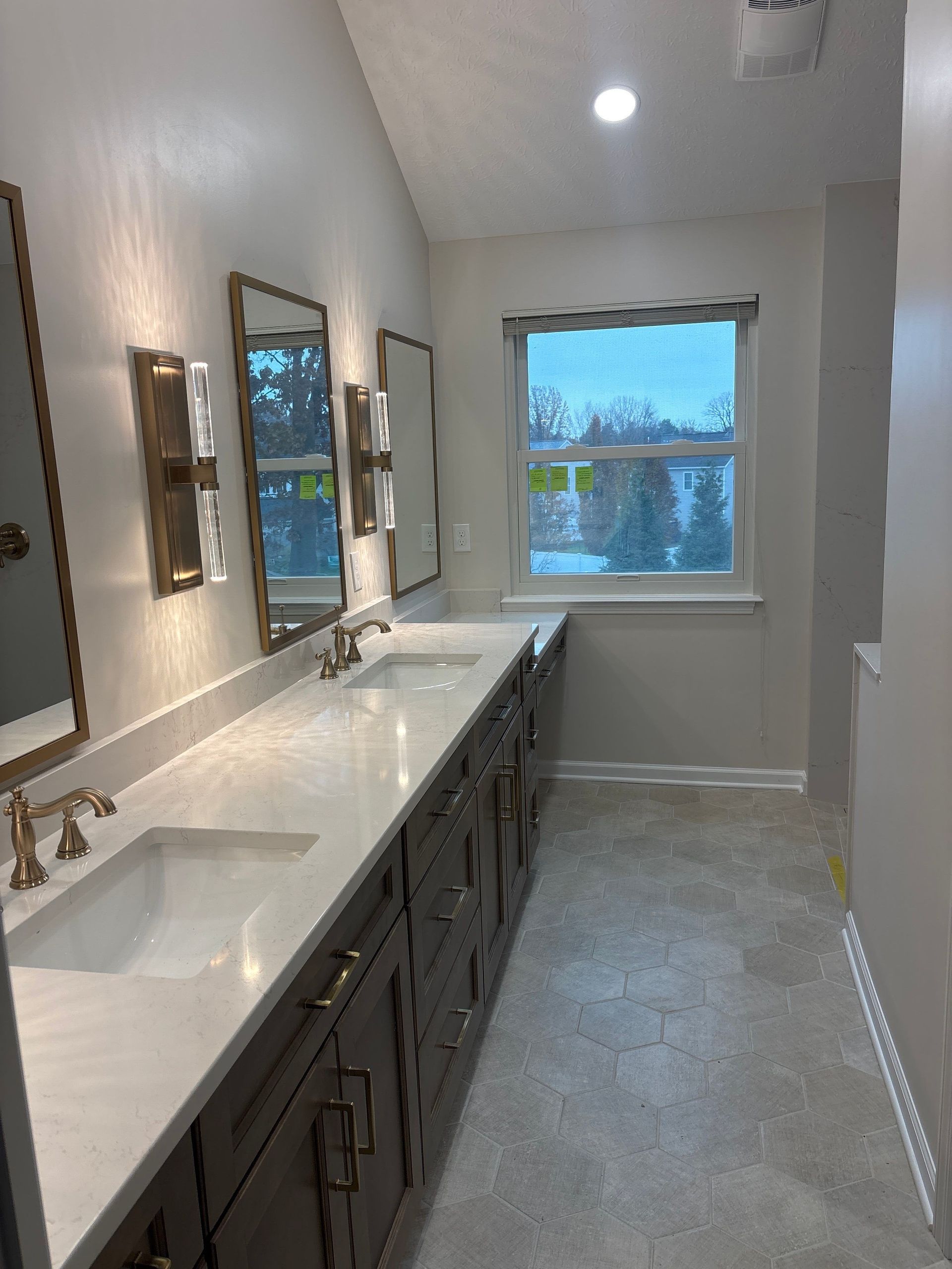 Long bathroom with white double vanity, dark cabinets, mirrors, and a window at the far end.