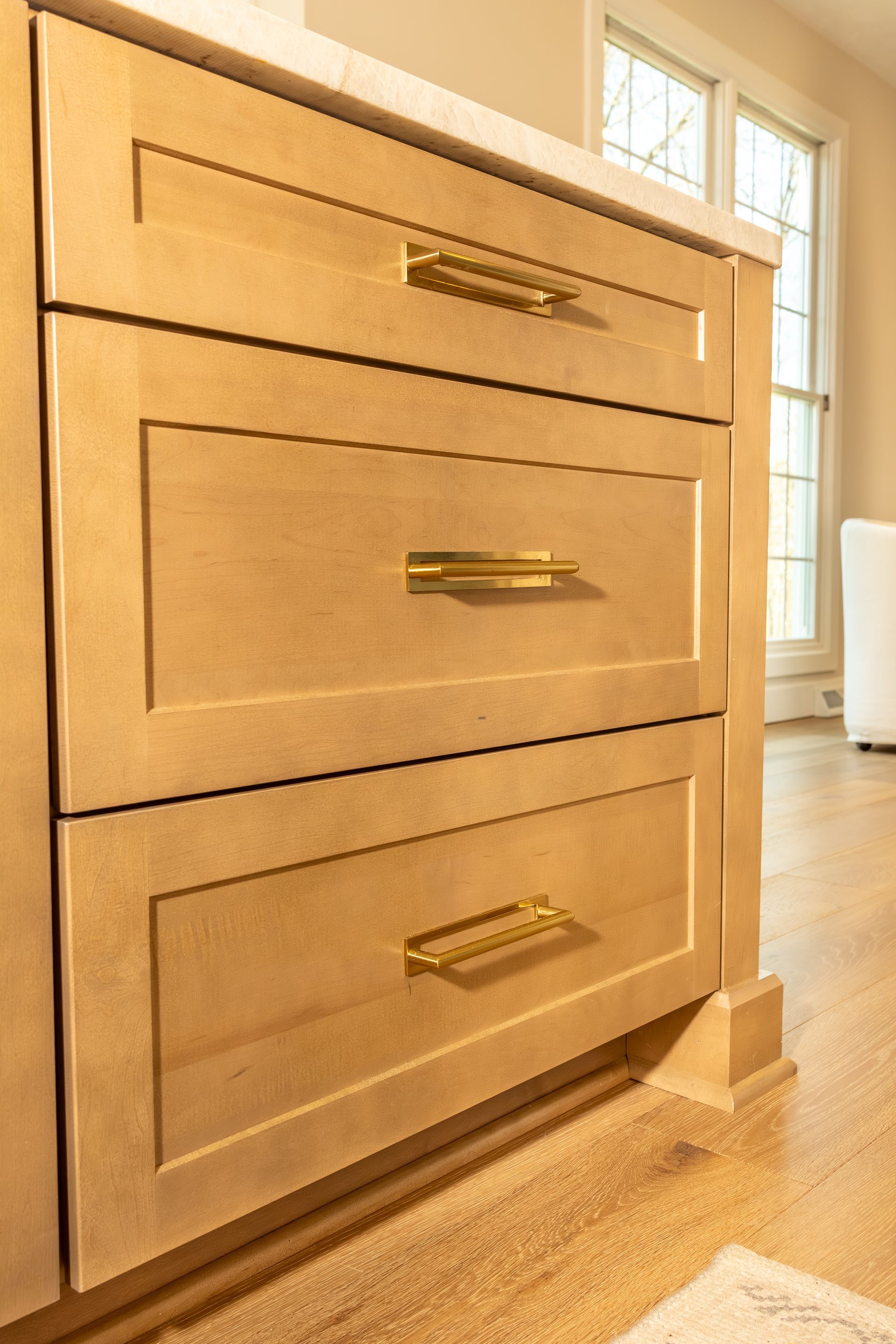Light wood kitchen cabinets with gold handles beside a bright window on a hardwood floor