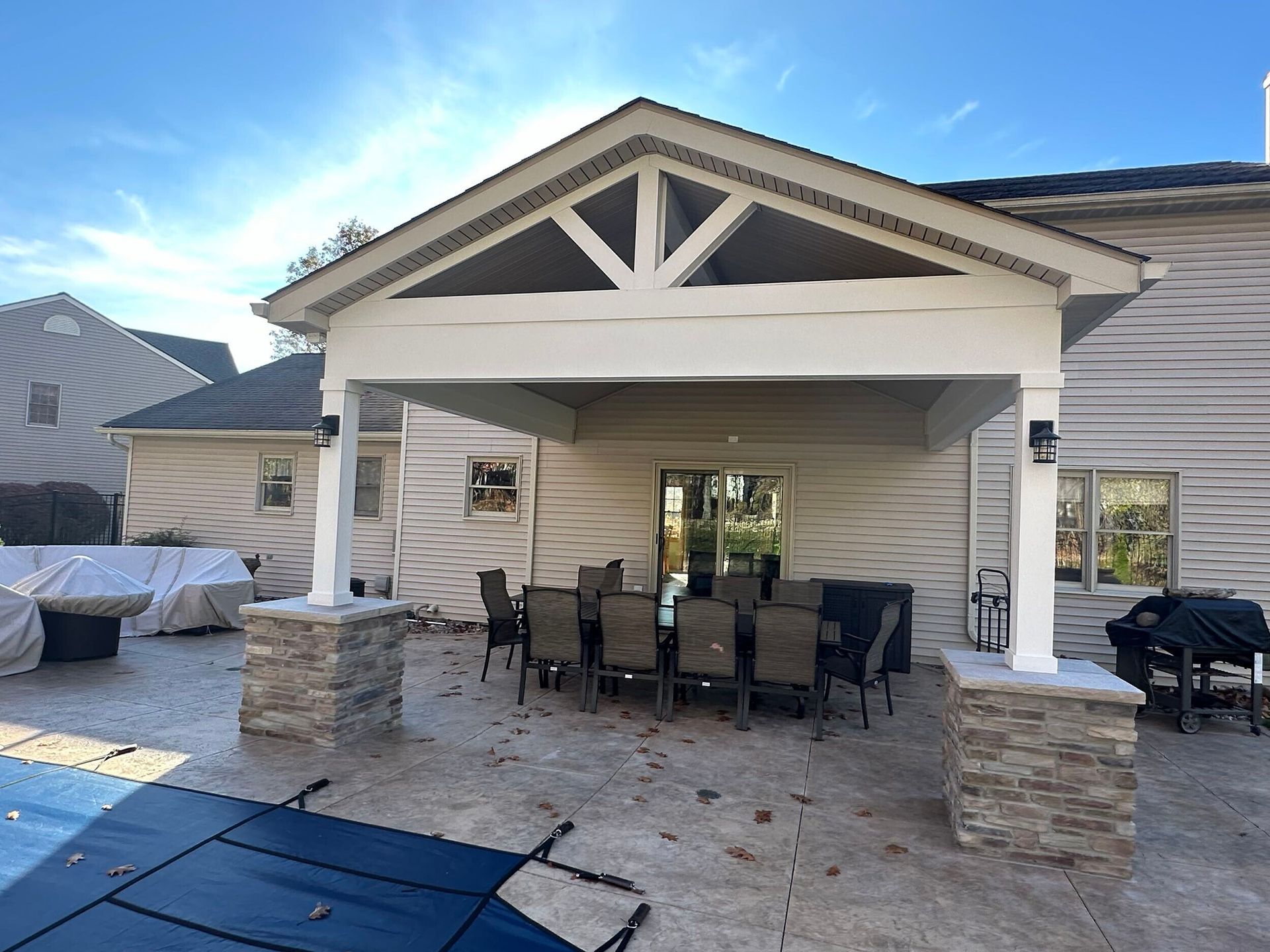 Covered patio with dining table and chairs outside a house. Stone columns and brown siding.