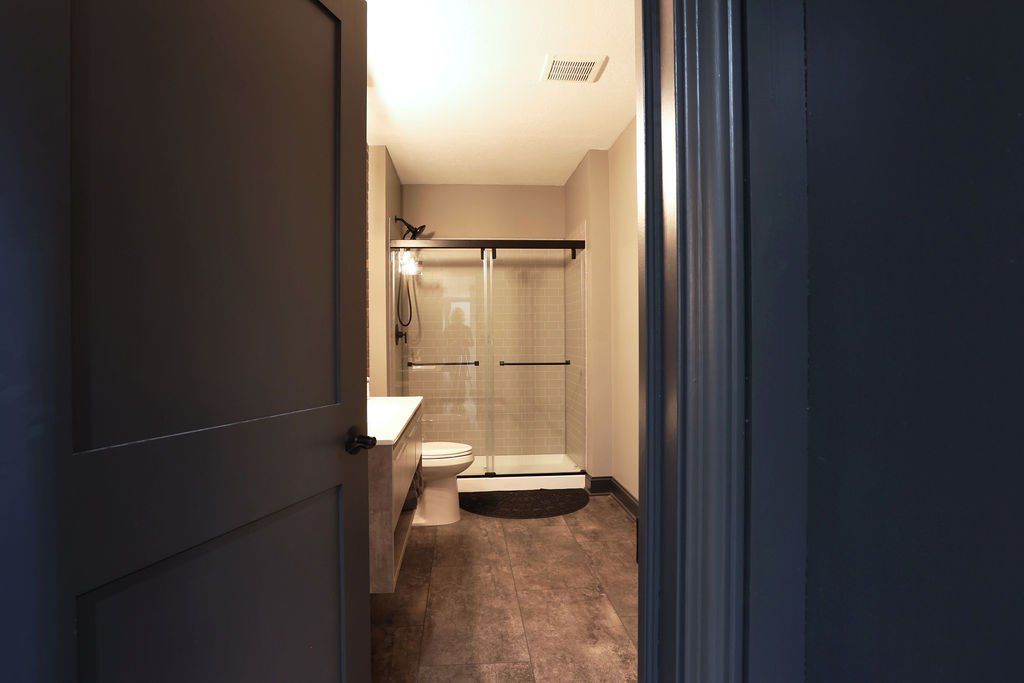 Bathroom interior seen through a partially open gray door; features a shower, toilet, and vanity.