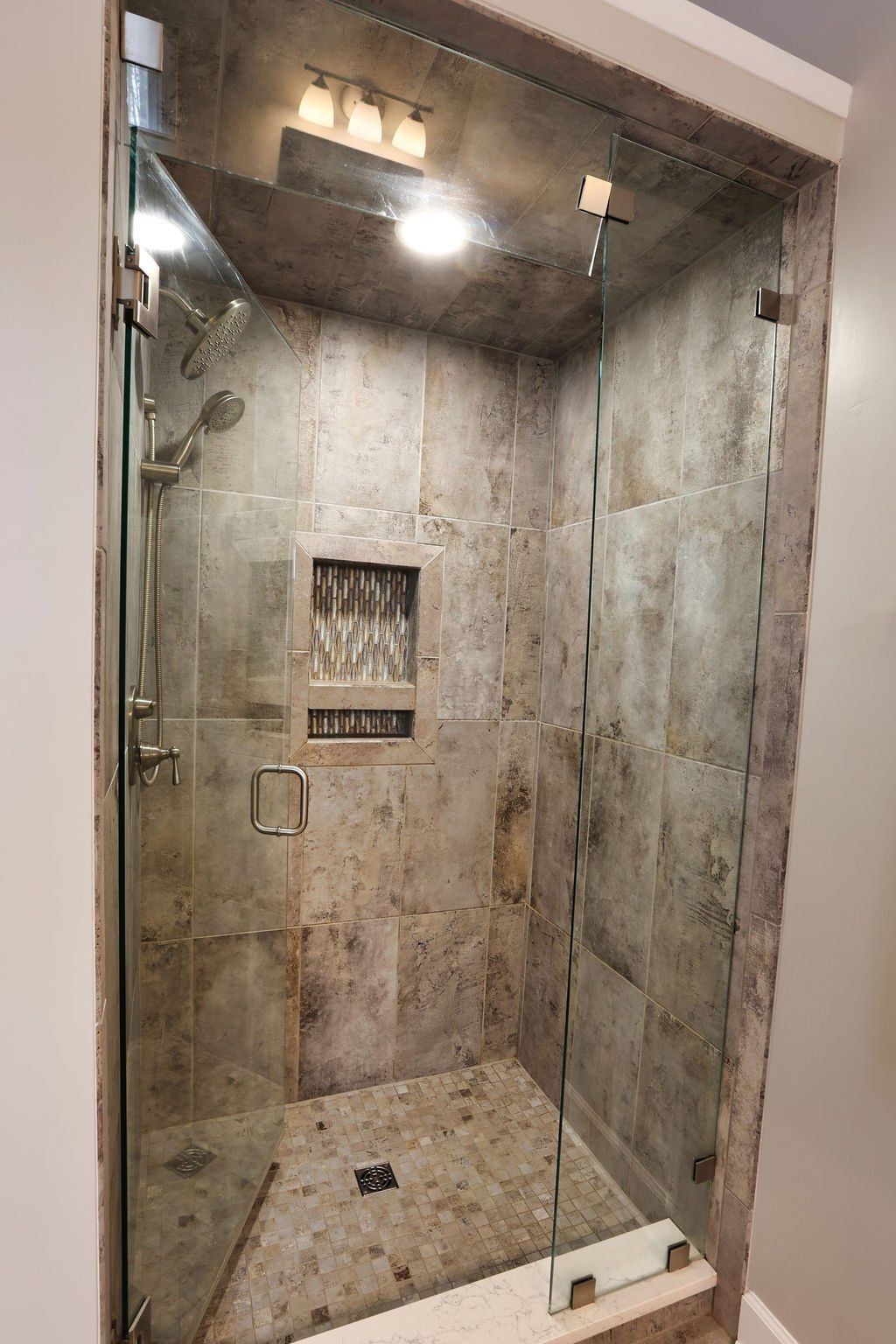 Shower with glass doors, gray tile walls, built-in shelf, and overhead lighting.