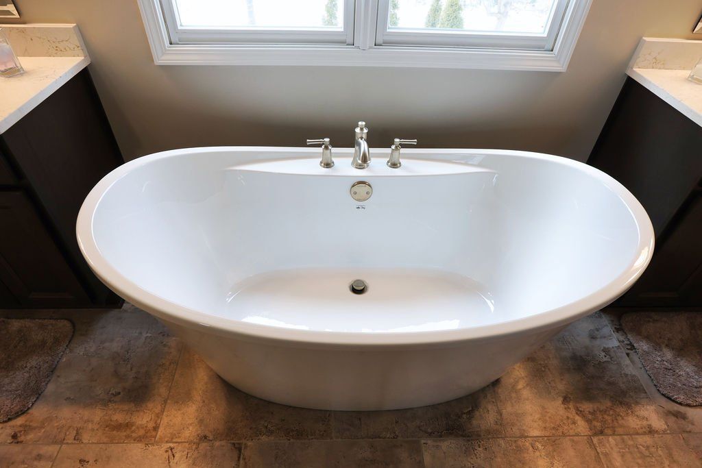Oval white bathtub in a bathroom, with faucet and view of window, next to dark cabinets.