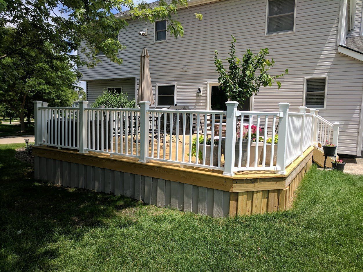 A white-railed deck with vertical supports, built around a two-story house, surrounded by grass.