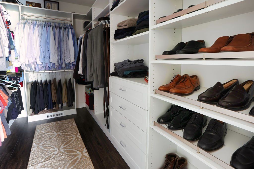 Walk-in closet with white shelves and drawers. Shoes and clothes are neatly organized. Dark wood floor and beige rug.