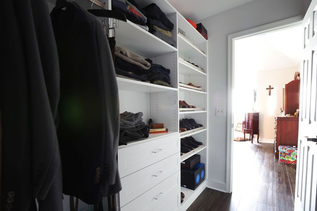 Closet with white shelving, clothes, shoes. Doorway leads to bedroom with furniture.