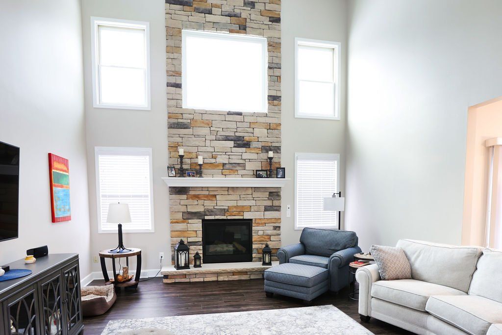 Living room with stone fireplace, high ceilings, windows, and furniture in neutral tones.