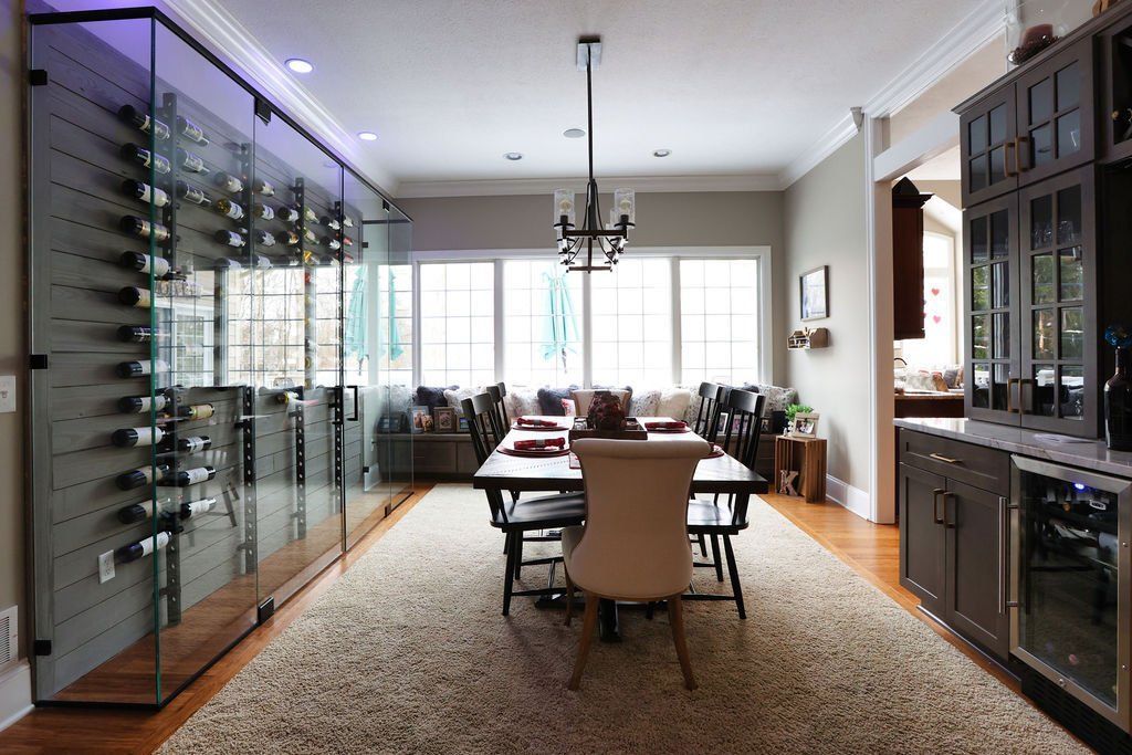 Dining room with a glass-enclosed wine rack, table, and windows, with a rug and dark cabinets.