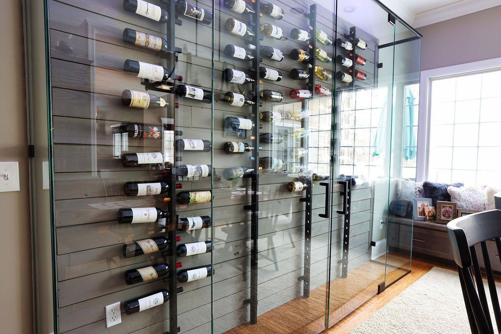 Wine bottles displayed in a glass-enclosed storage unit against a gray wall, near a window and dining area.