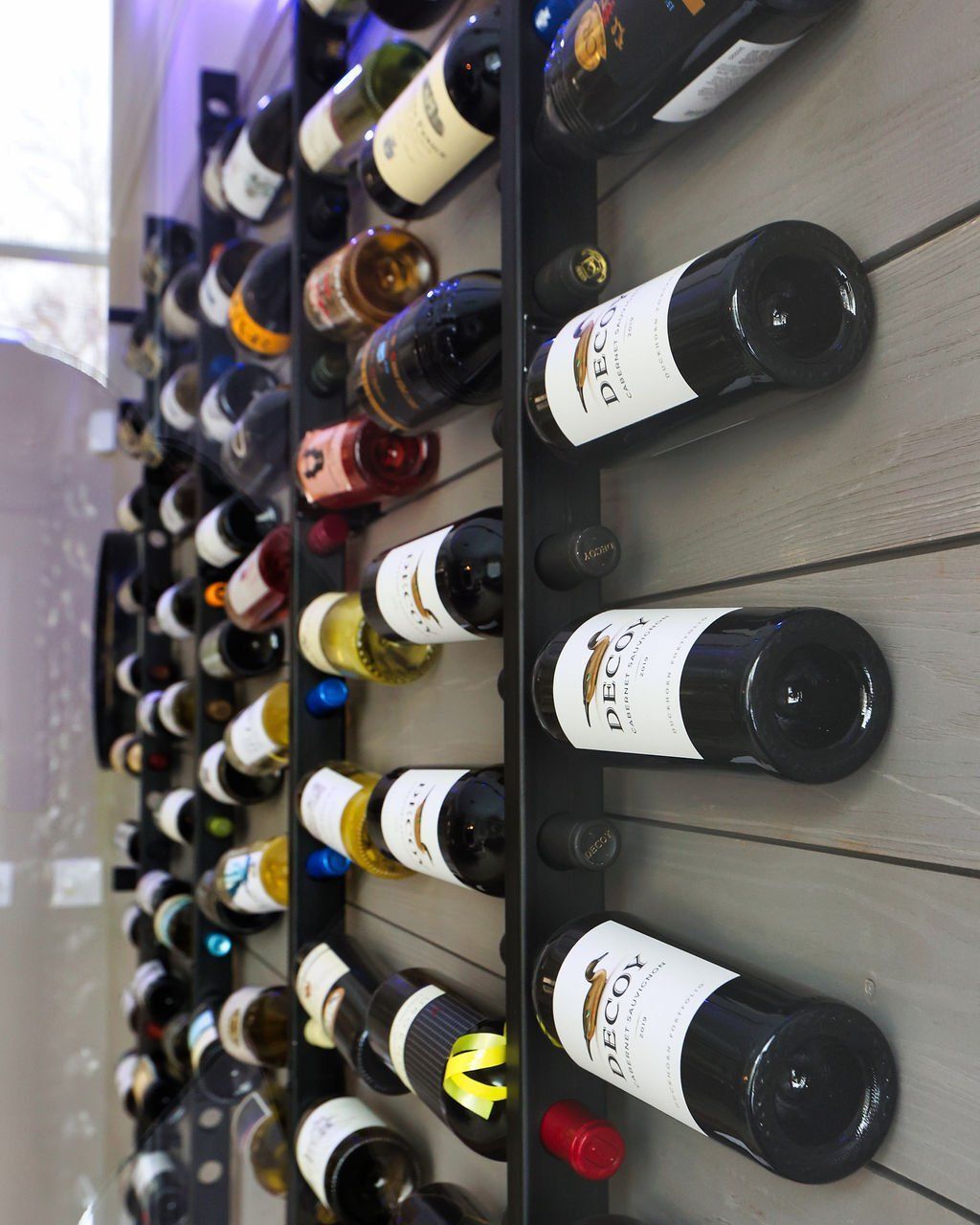 Wine bottles on a wall-mounted rack, arranged horizontally. Black metal against a light gray wood-paneled wall.