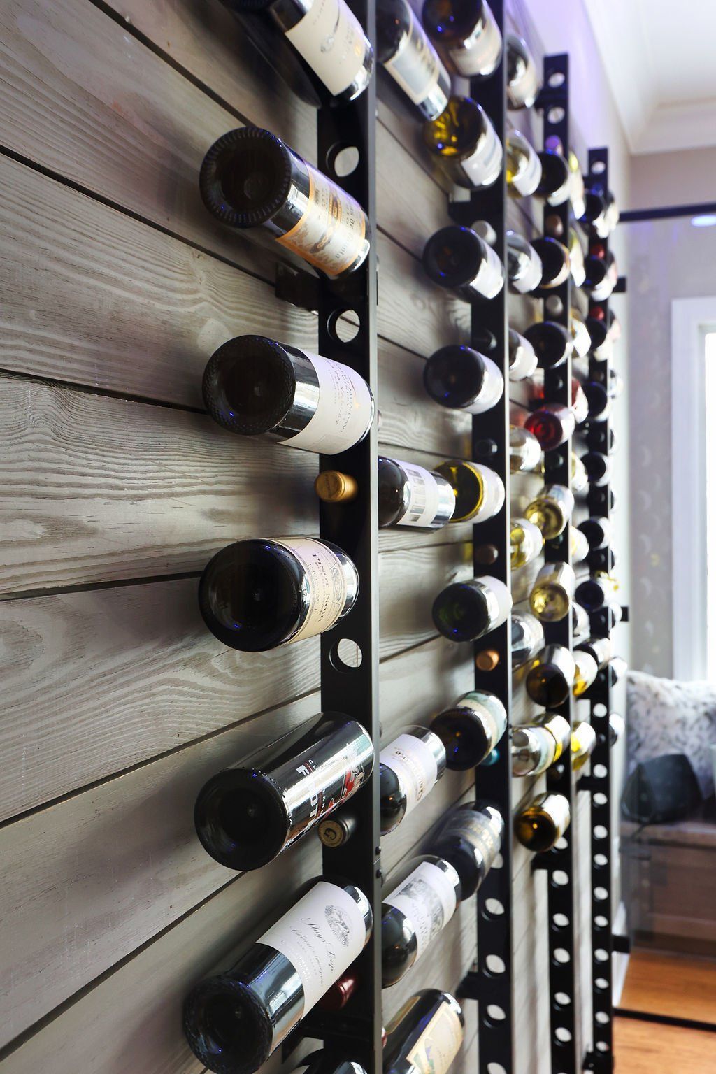 Wall-mounted black wine racks with full and empty bottles against gray-washed wood paneling.