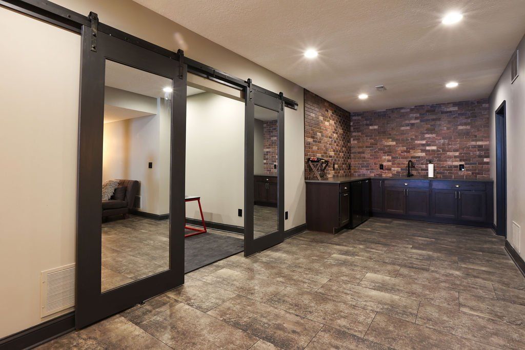 Basement gym area with mirrored sliding doors, brick wall, dark cabinets, and carpet flooring.