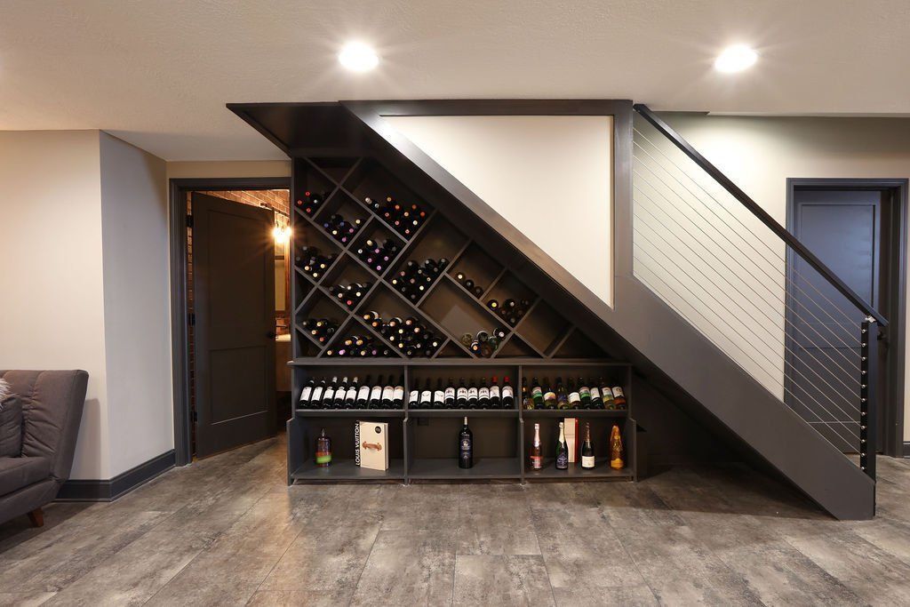 Wine rack built under a staircase, with dark shelves filled with bottles, in a modern basement setting.