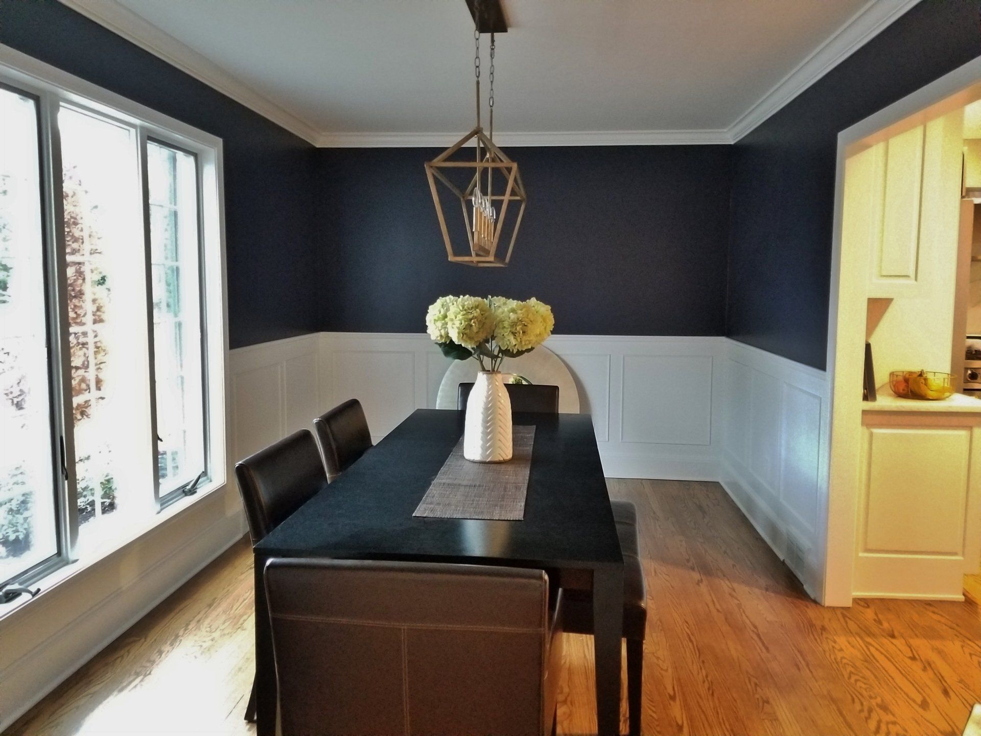 Formal dining room: dark blue walls, white wainscoting, black table, gold light, white flowers.