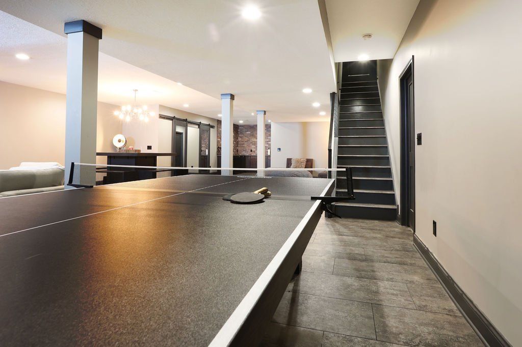 Spacious basement with a pool table, stairs, and supporting columns. Neutral color scheme, good lighting.