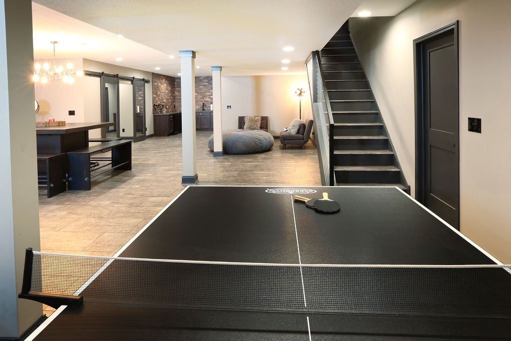 Ping pong table in a finished basement with stairs and a lounge area in the background.
