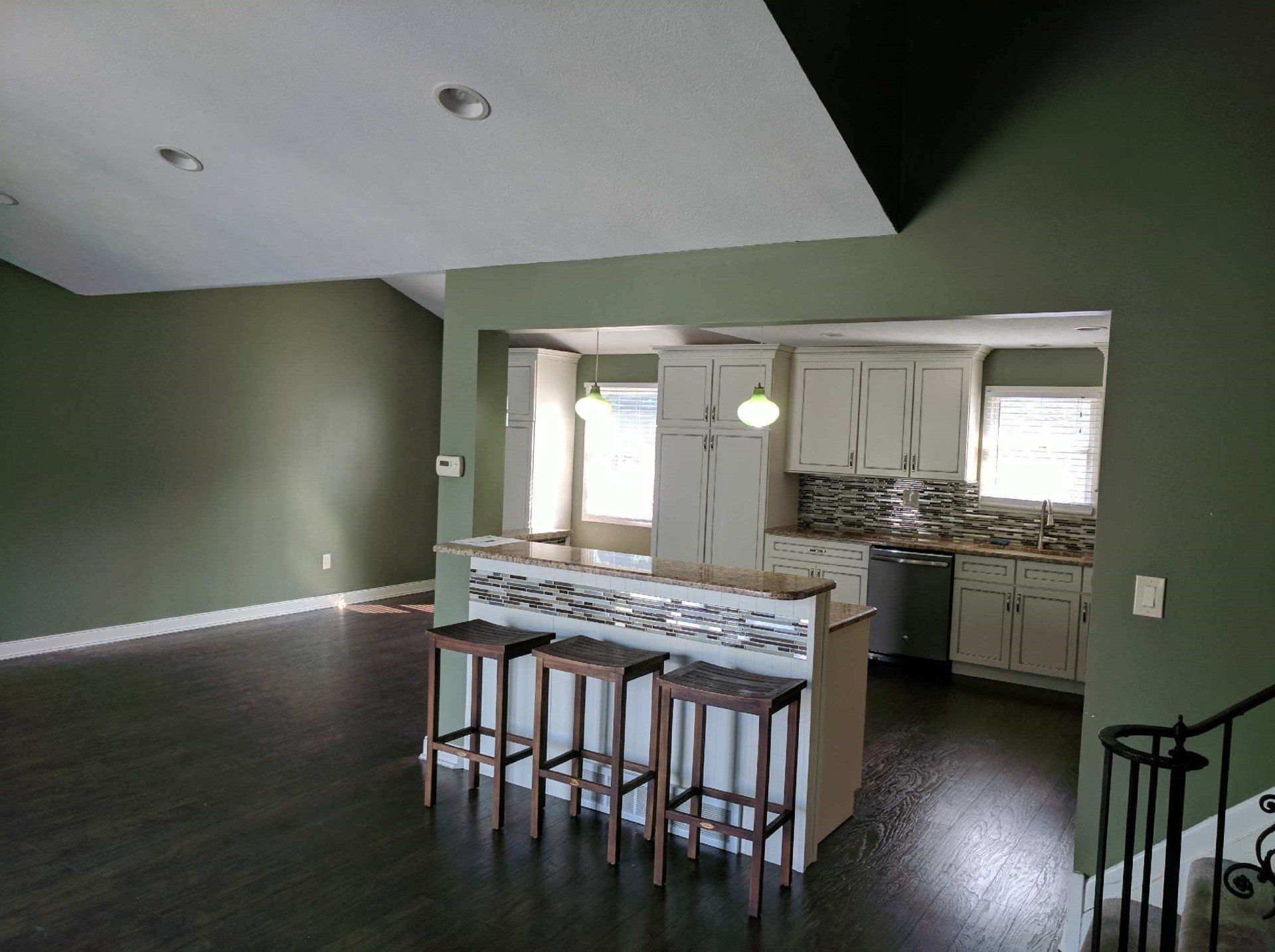 Kitchen and living area with a dark floor, white cabinets, and green walls. A bar with three stools separates the two spaces.