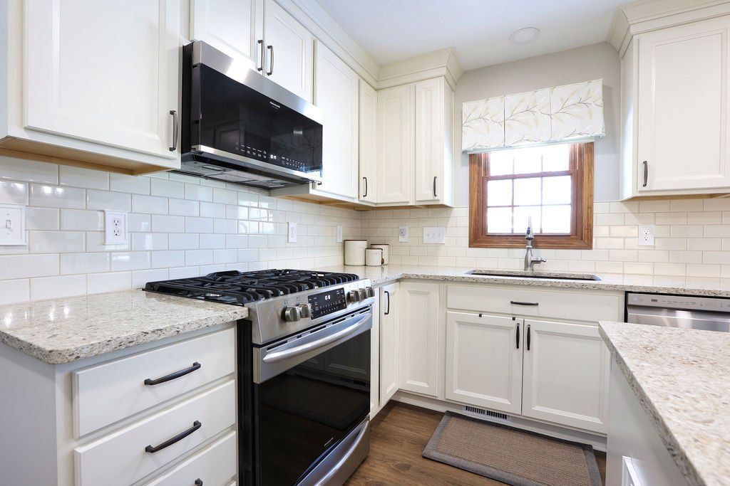 Bright white kitchen with granite counters, gas stove, black microwave, and window above the sink.