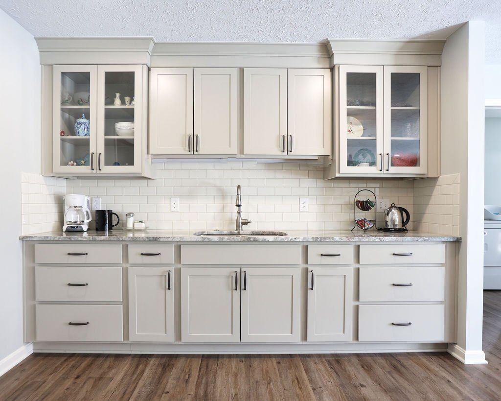 Modern kitchen with light gray cabinets, glass-front uppers, white tile backsplash, and dark wood floor.