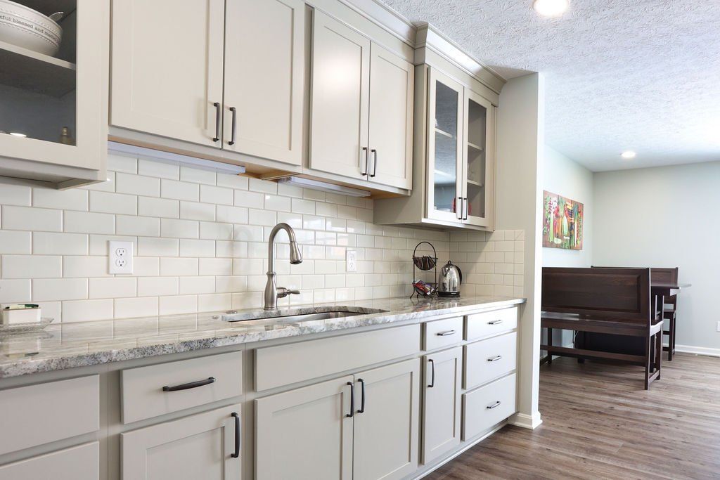 Kitchen with white cabinets, light backsplash, and a view into a dining area with a dark wooden bench.
